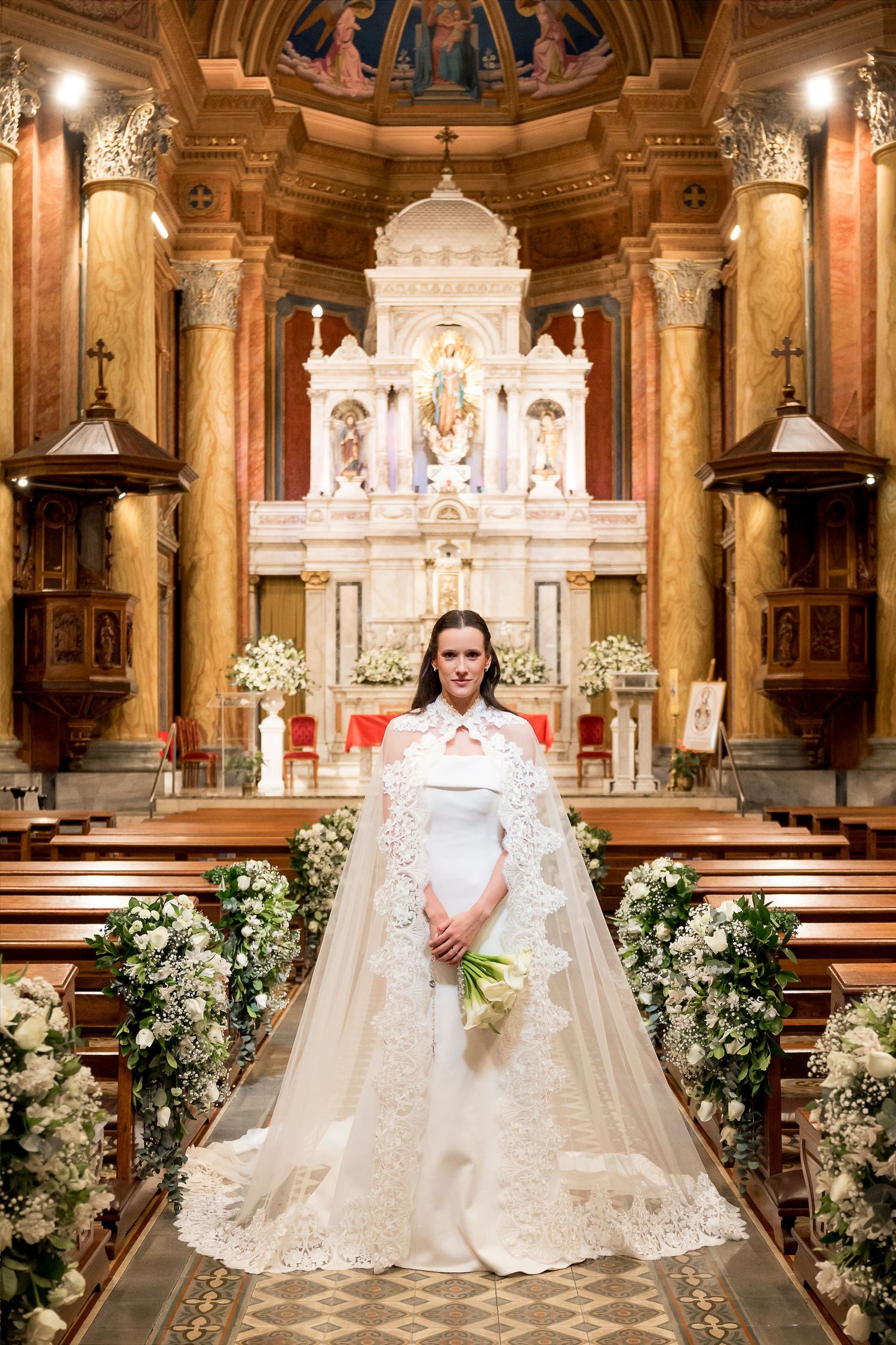 Bride in white lace cape gown holds bouquet at ornate church altar adorned with flowers.