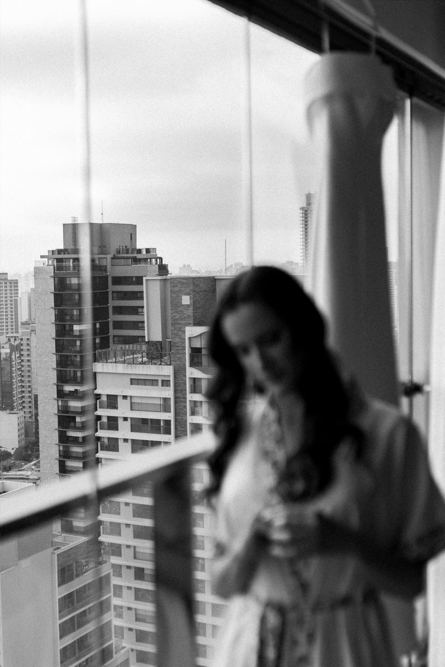 Black and white photo of a woman in white dress standing by a window with city skyline buildings in background.