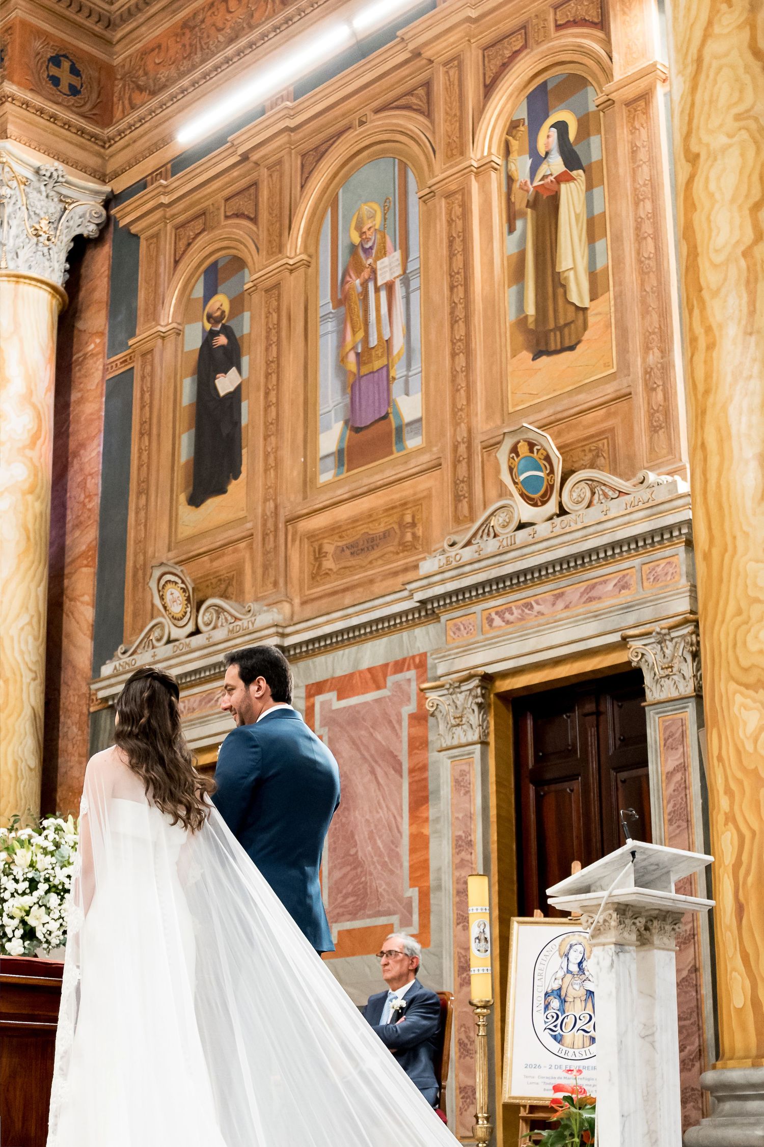 Bride and groom stand before ornate frescoed altar in elegant Italian church during wedding ceremony.