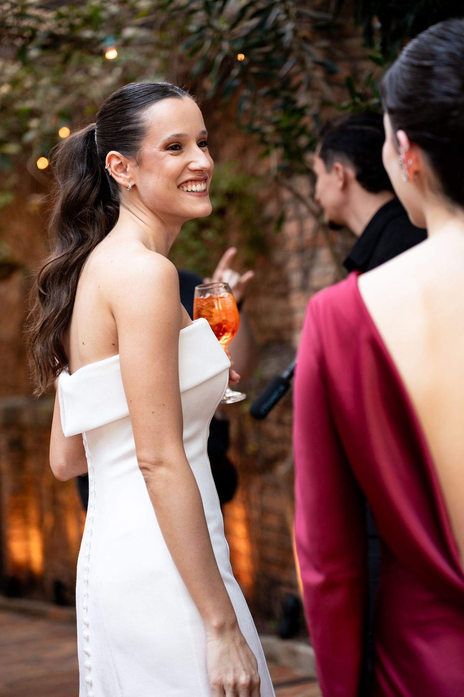 Smiling woman in white off-shoulder dress holding cocktail at elegant outdoor wedding reception.