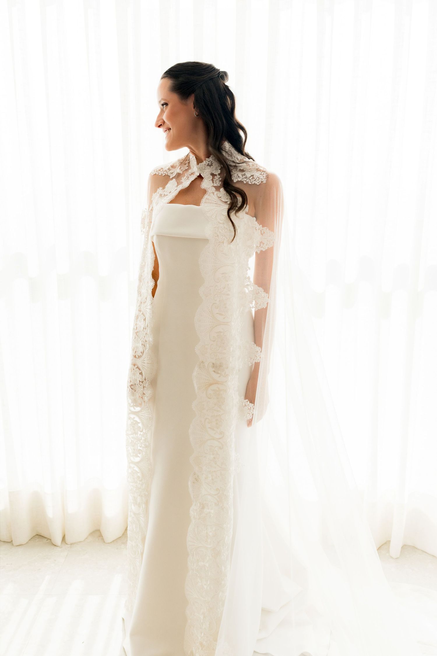 Bride in elegant white sheath gown with lace-trimmed veil standing by bright window, dark hair styled up.