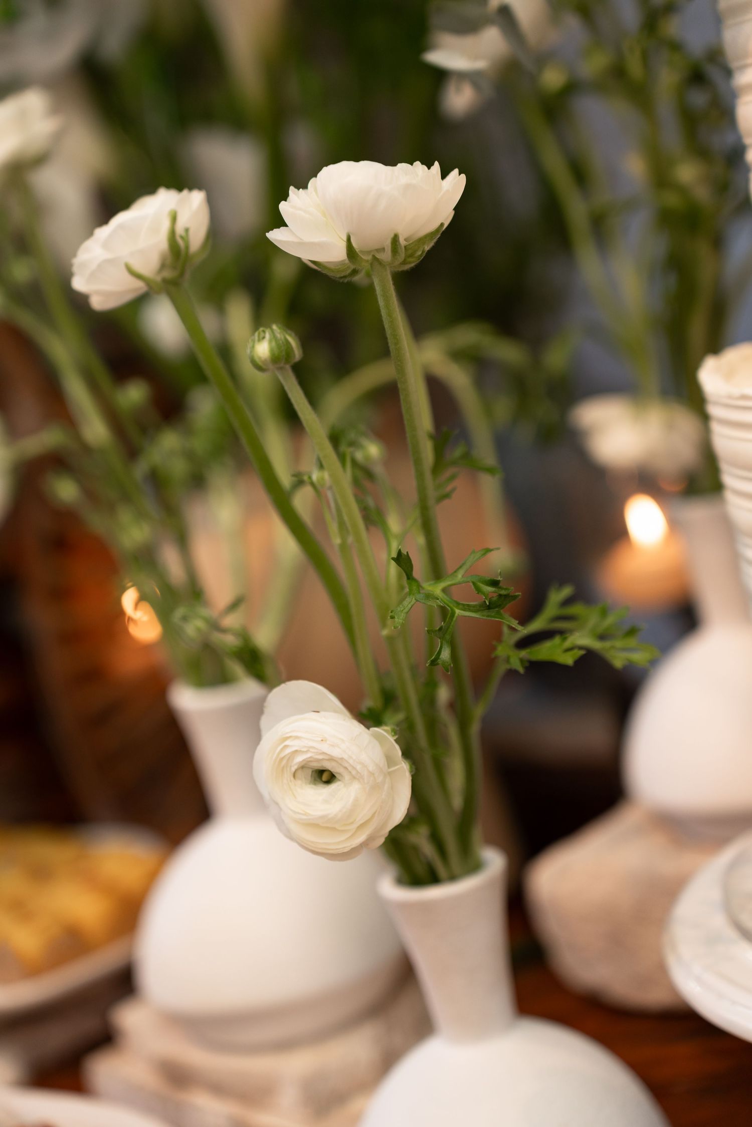 White ranunculus flowers in minimalist white ceramic vases on a decorated table with soft candlelight.