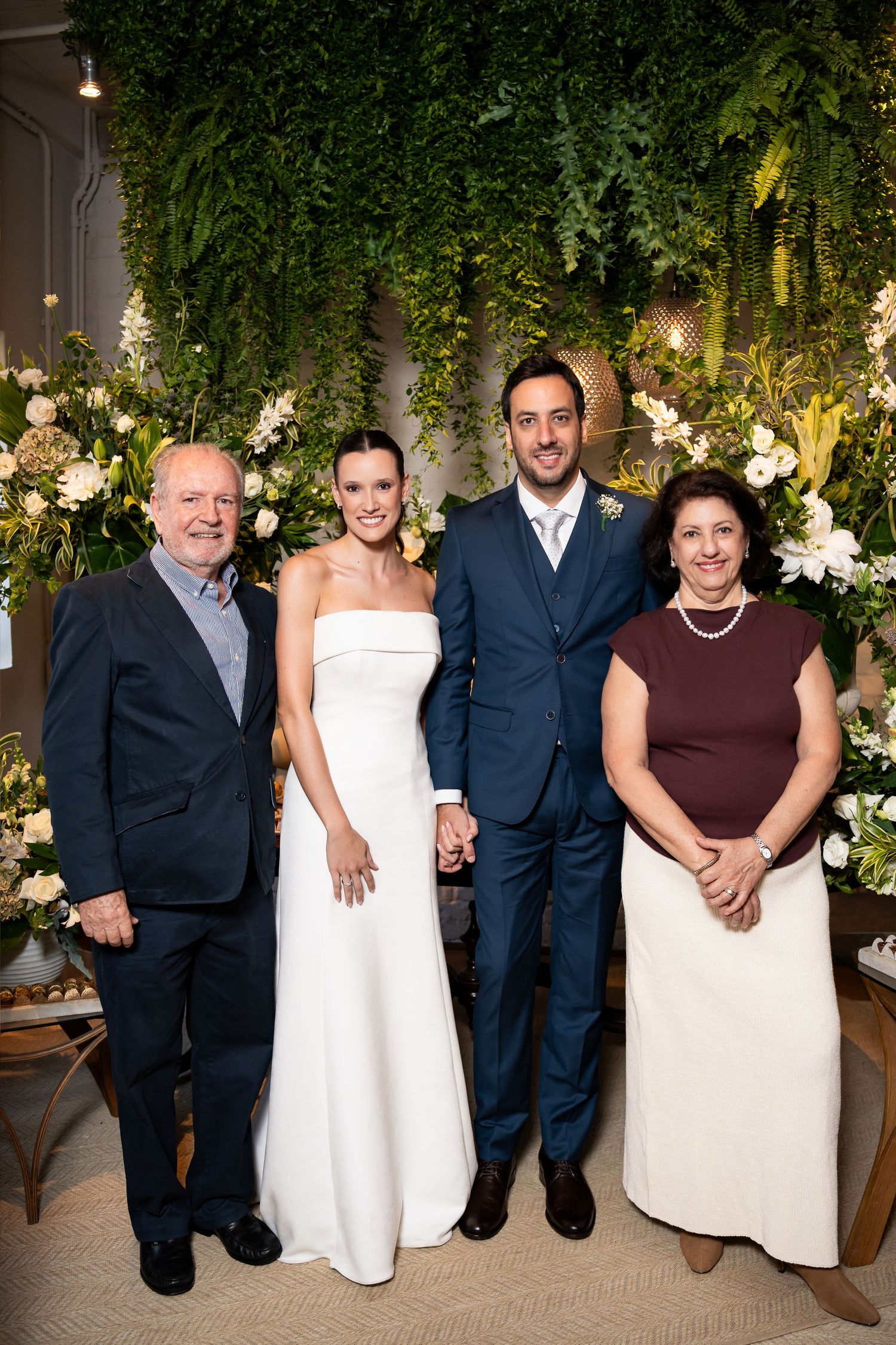 Wedding couple in white gown and navy suit pose with family before lush green floral backdrop.