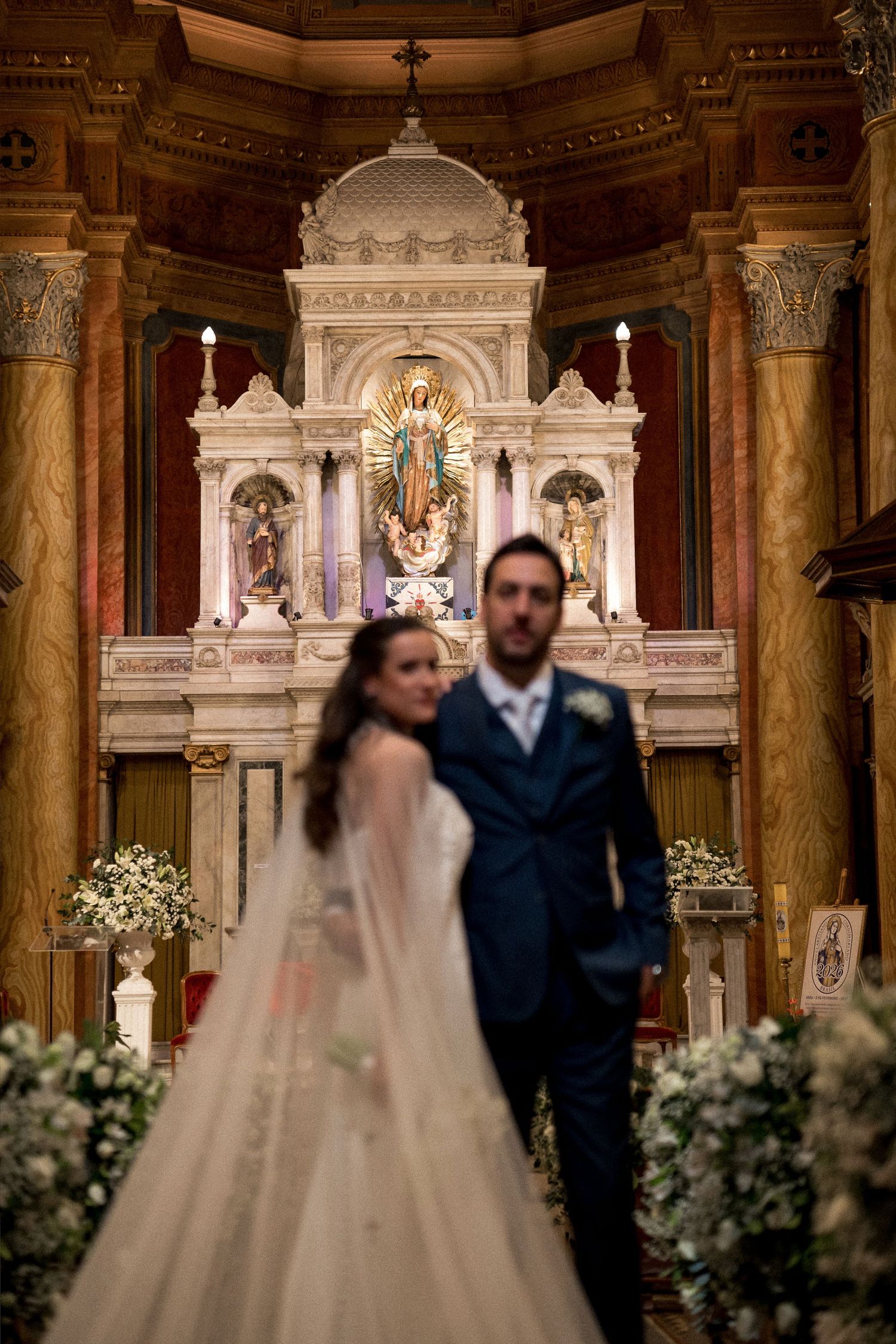Bride and groom standing before an ornate church altar adorned with flowers and a golden Madonna statue.
