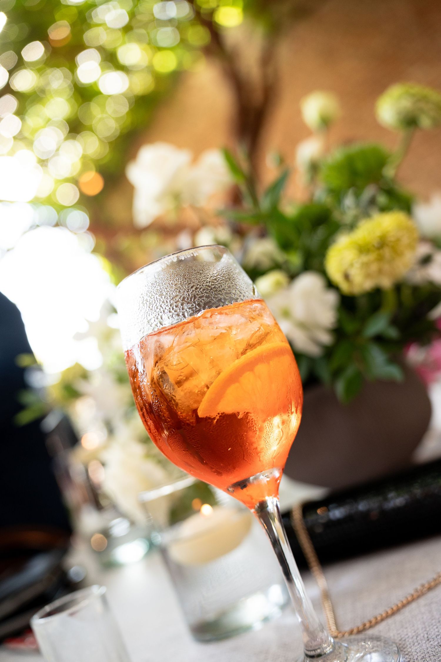A chilled Aperol Spritz cocktail with orange slice in a wine glass, surrounded by white flowers at an outdoor event.