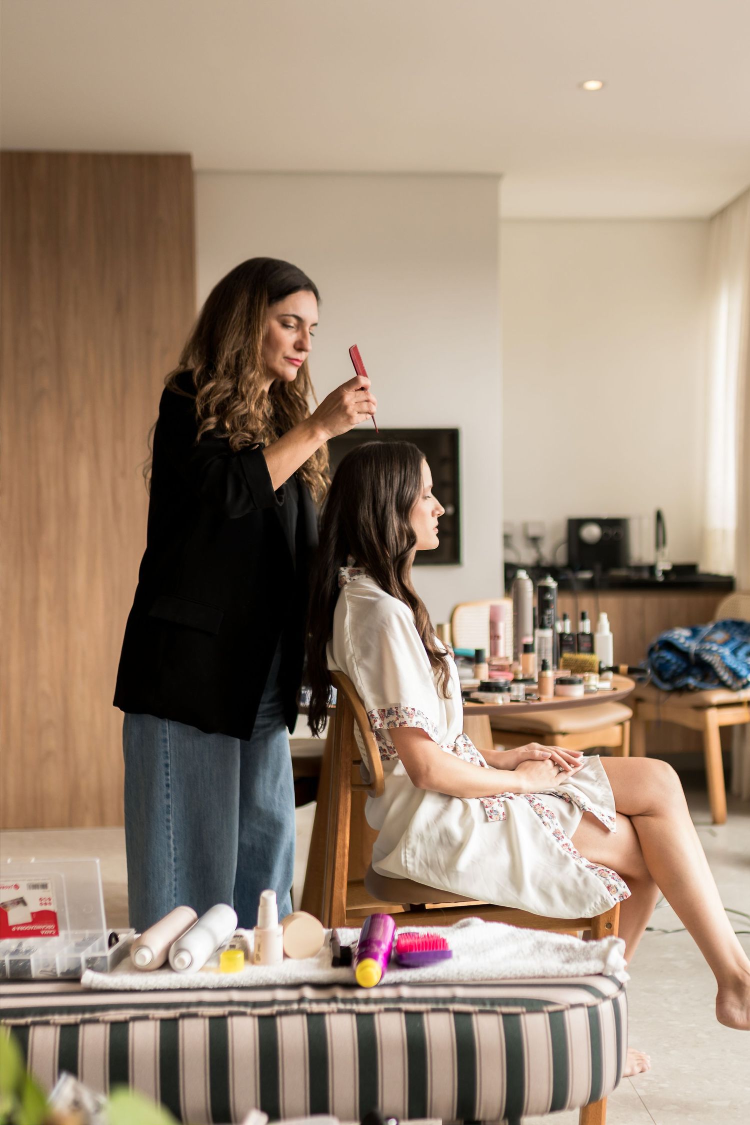A hairstylist in a black blazer styles a seated woman's hair in a bright modern room with makeup products nearby.