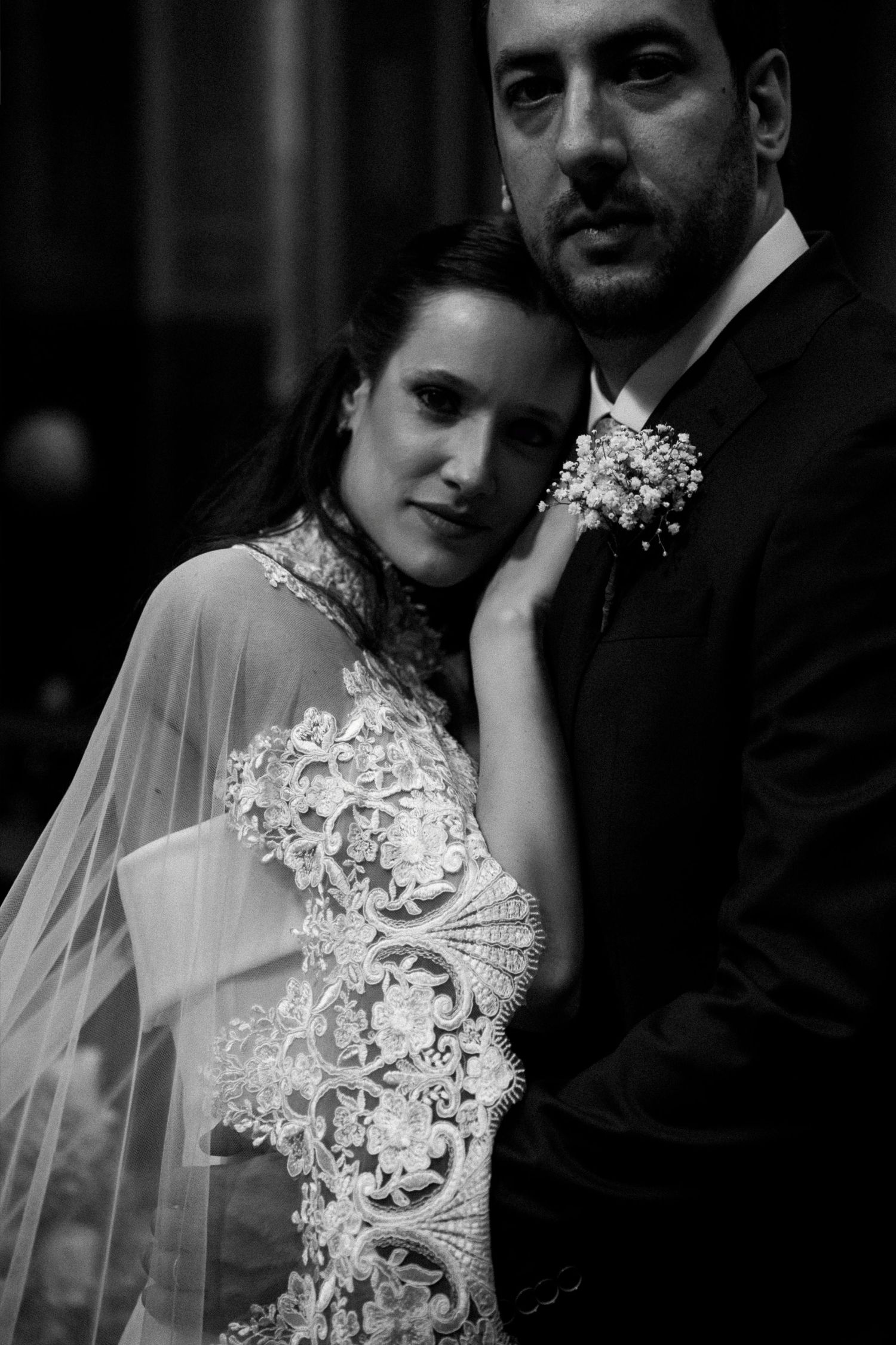 Black and white wedding portrait of bride in lace gown with veil leaning against groom in suit with boutonniere.