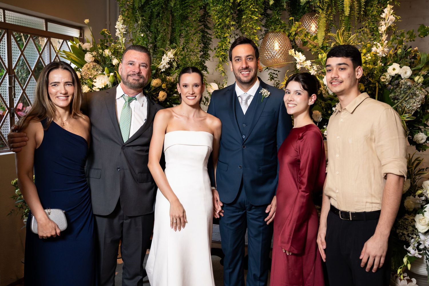 Wedding group photo with bride in white dress, groom in navy suit, and four guests posing before floral backdrop.