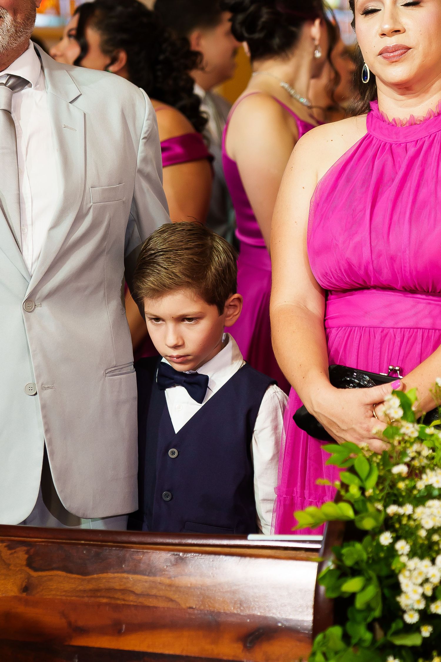 Young boy in navy vest and bow tie stands solemnly at a wedding ceremony beside adults in formal attire.
