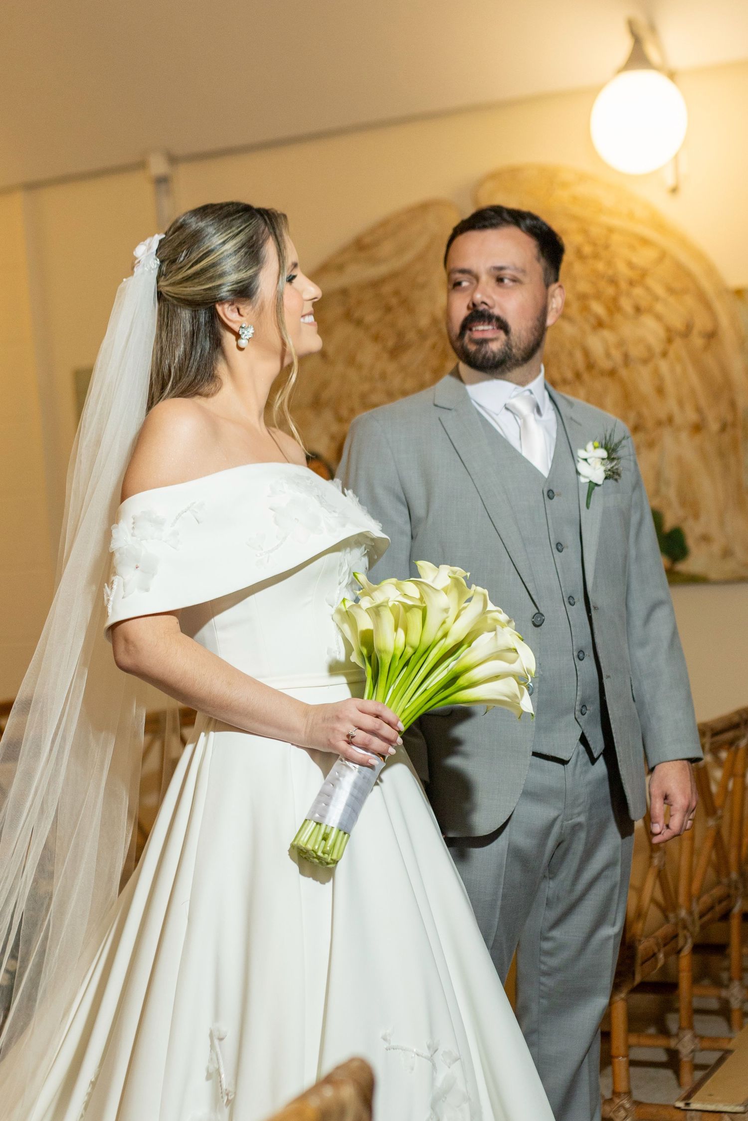 Bride in white off-shoulder gown holding calla lily bouquet stands with groom in gray suit at elegant wedding ceremony.