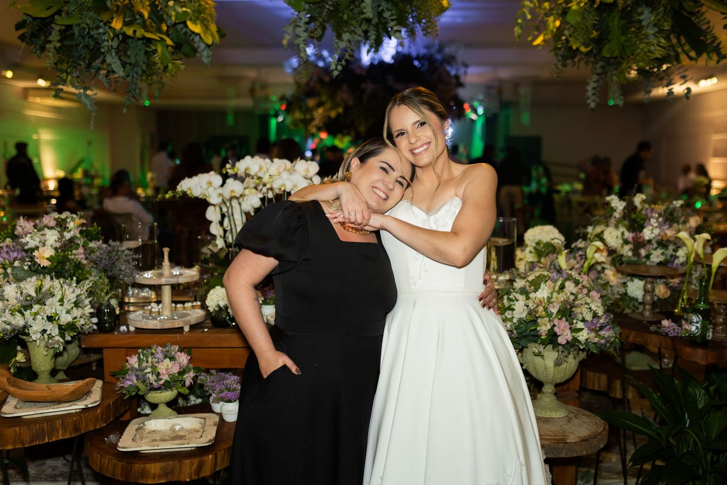 Two women pose together at an elegant wedding reception with floral arrangements and green ambient lighting.