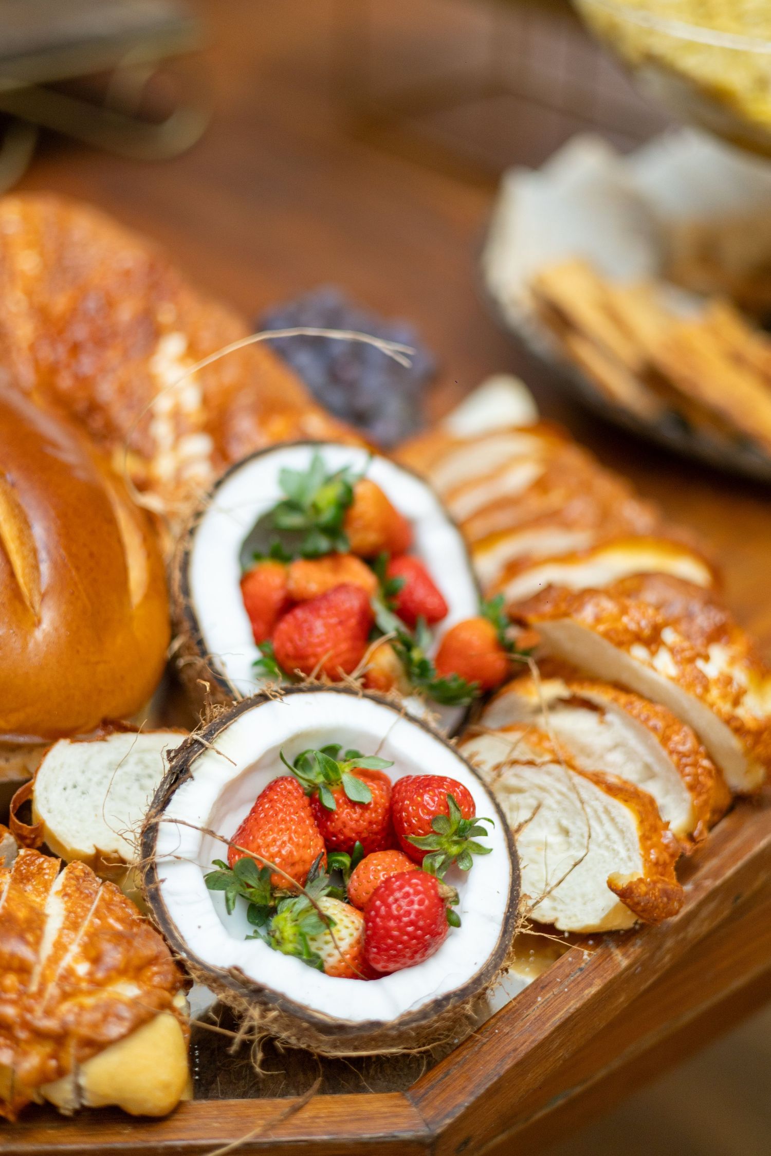 Fresh strawberries served in coconut halves on a wooden board with assorted breads and pastries.