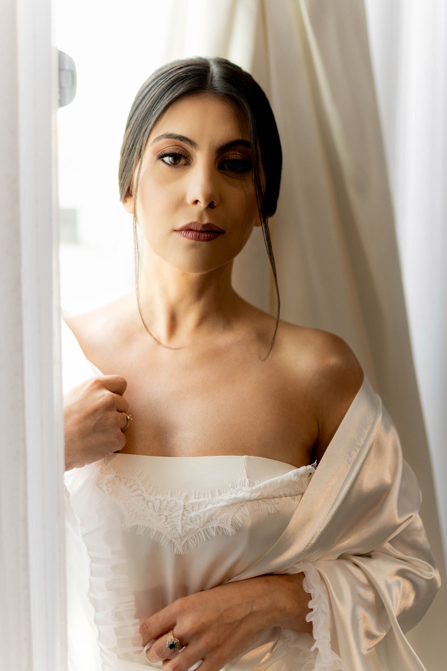 Elegant bride in white lace slip and satin robe posing near bright window with ring.