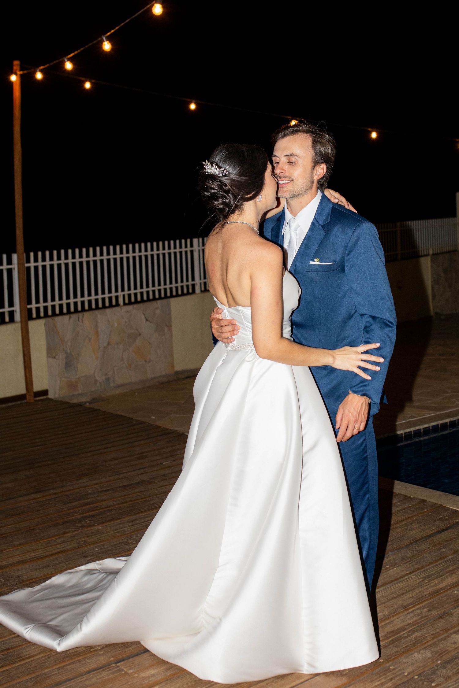 Bride and groom share first dance outdoors at night under string lights near a pool, bride in white gown, groom in blue suit.
