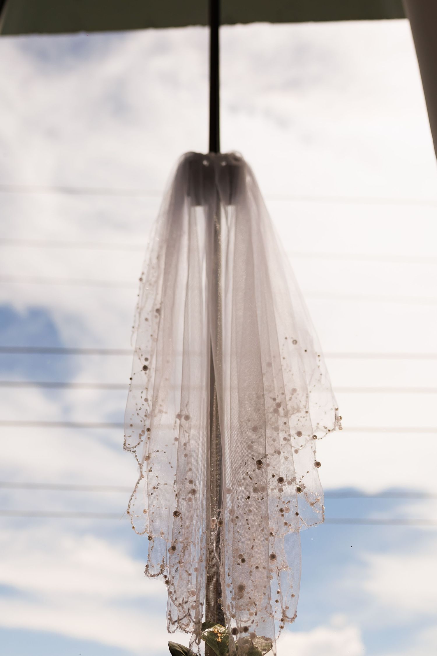 Elegant white bridal veil with gold beaded details hanging against a blue sky backdrop.