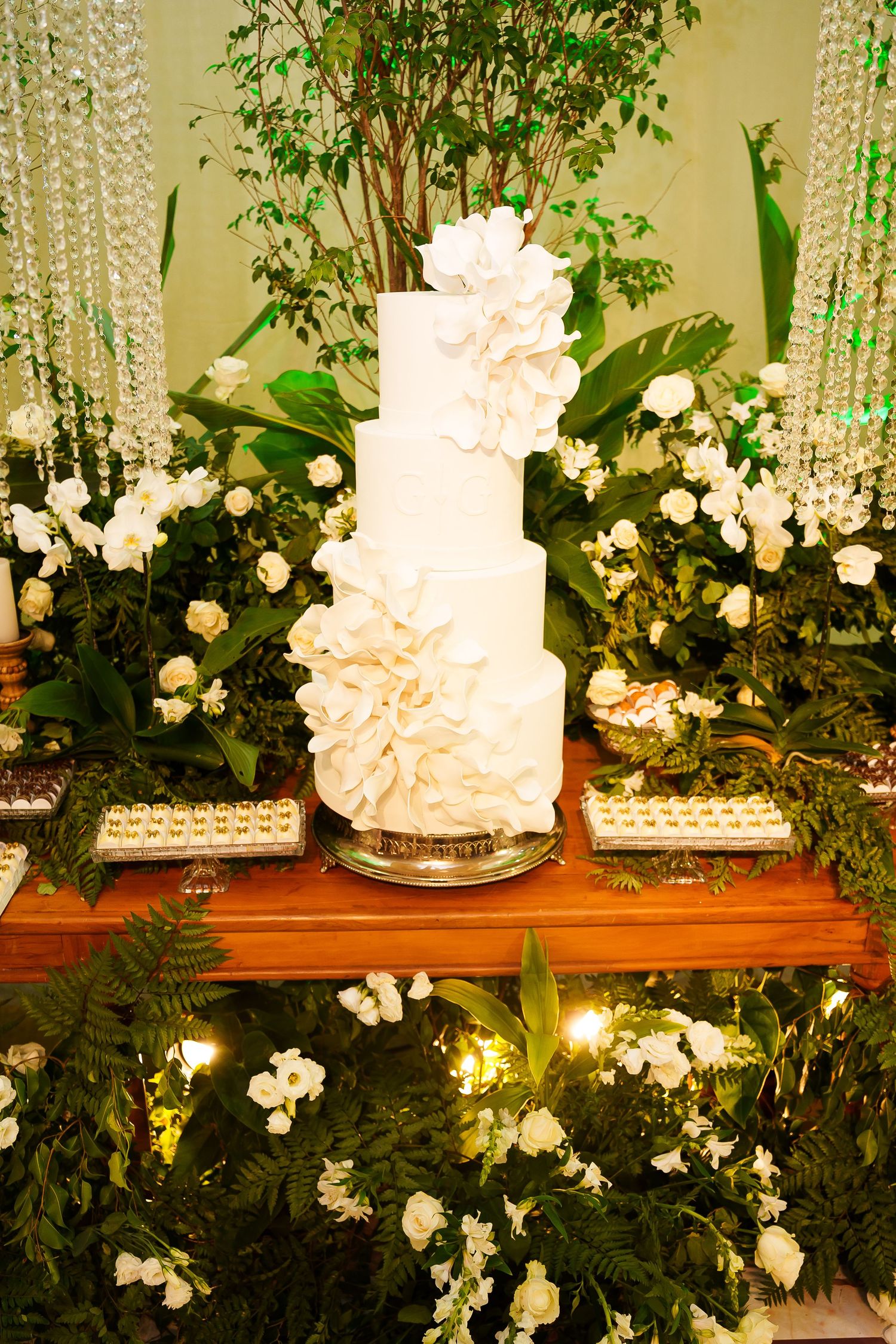 Elegant white tiered wedding cake adorned with floral details, surrounded by lush greenery and white flowers on a wooden table.