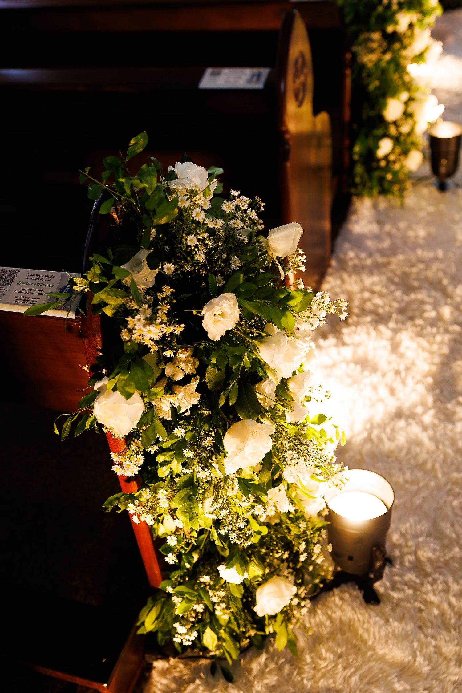 White floral wedding aisle decor with greenery and candles adorning wooden church pews.