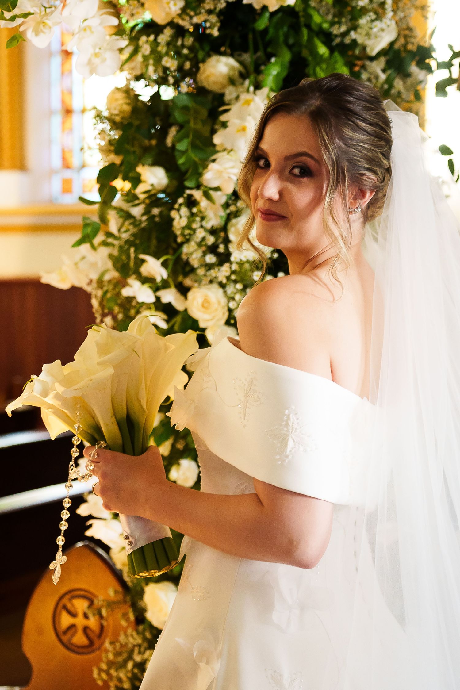 Elegant bride in off-shoulder white gown holding calla lily bouquet, wearing veil, posing before floral arch in church.