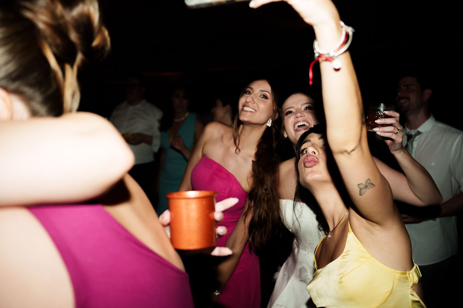 Women dancing and celebrating at a party, holding drinks, wearing colorful dresses in a dimly lit venue.