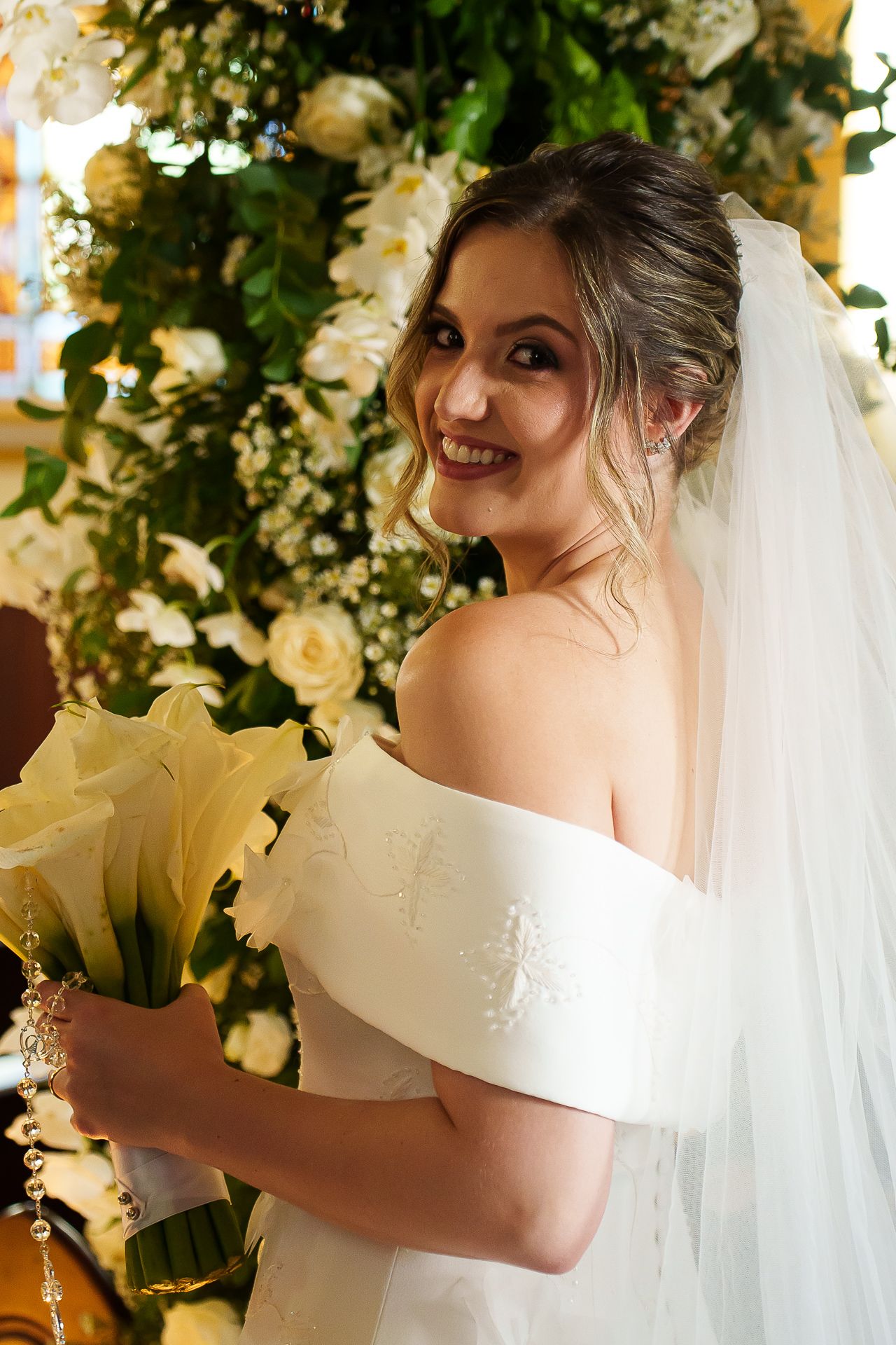 Smiling bride in off-shoulder white gown and veil holding yellow lilies bouquet near white floral arrangement.