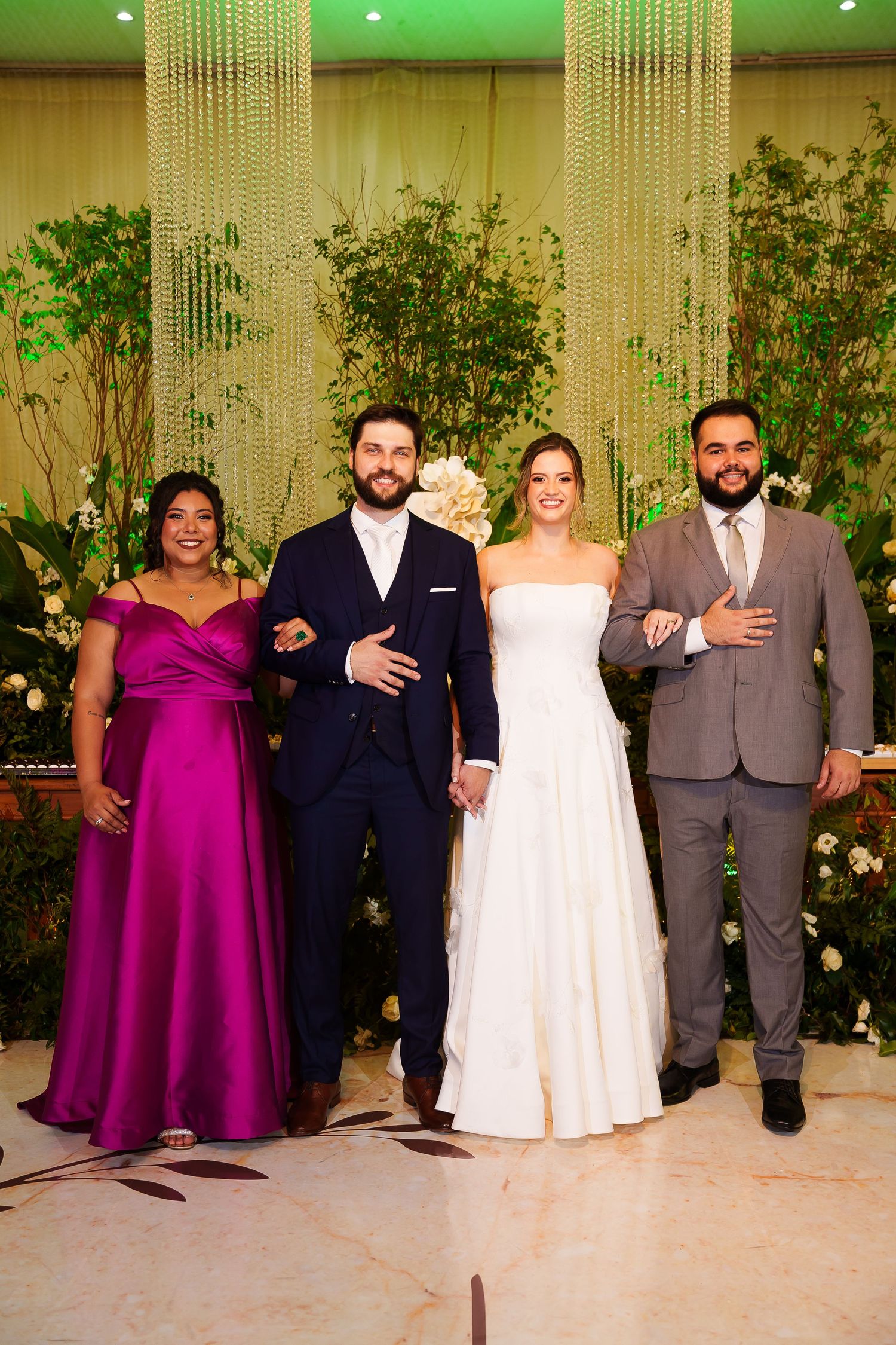 Bride and groom pose with two guests at elegant wedding reception with lush green floral backdrop.