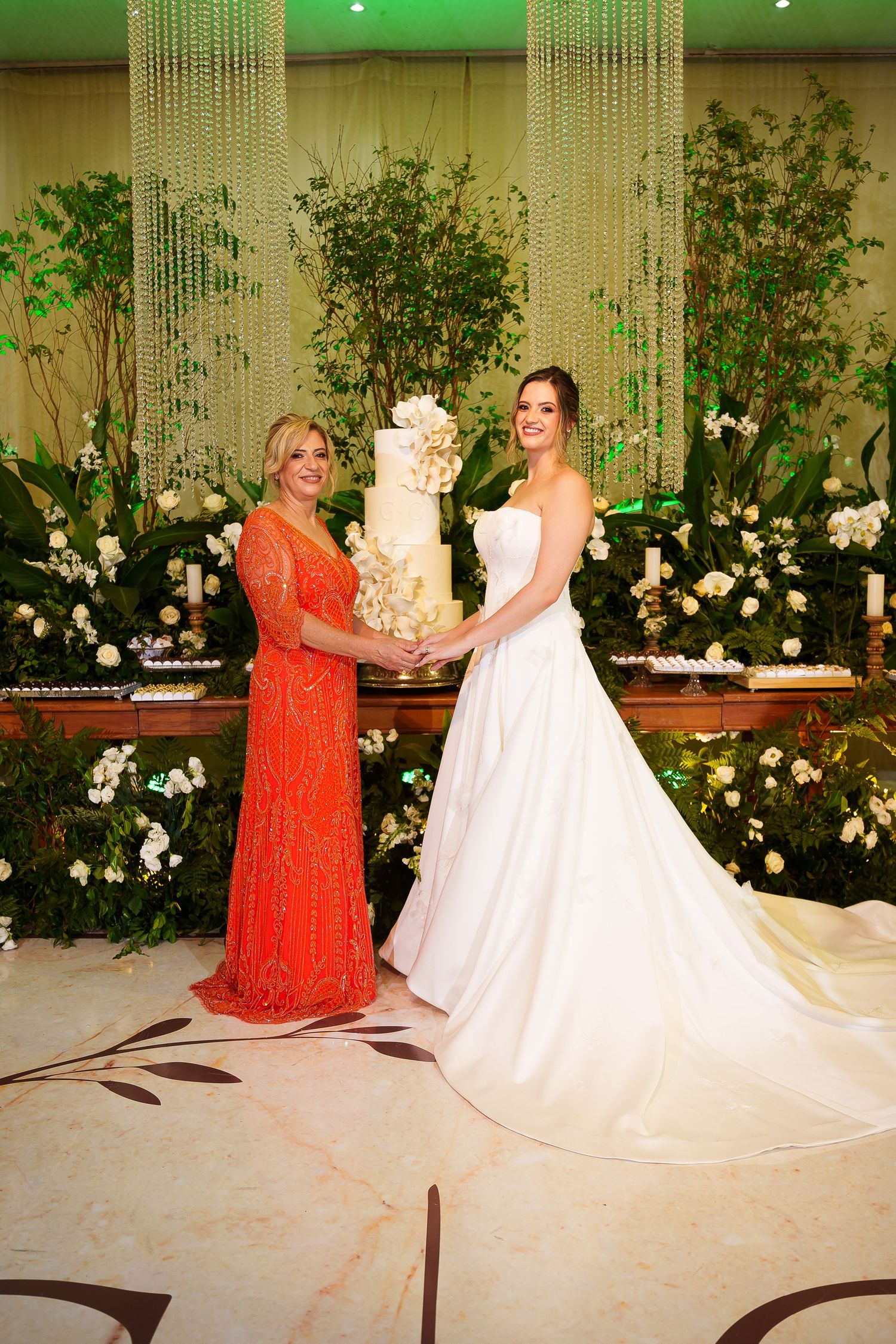Bride in white gown and woman in orange dress holding a white wedding cake at an elegant floral reception.