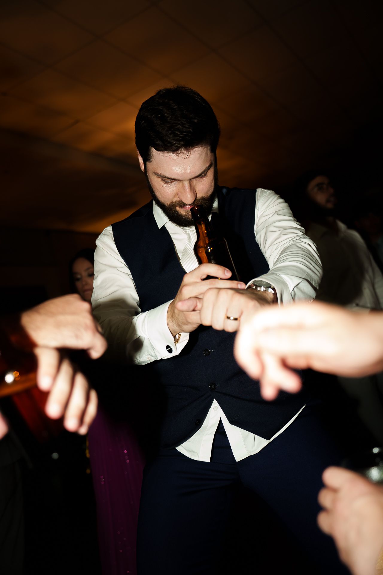 A bearded man in a vest dances energetically at a party, holding a beer bottle while guests point at him.