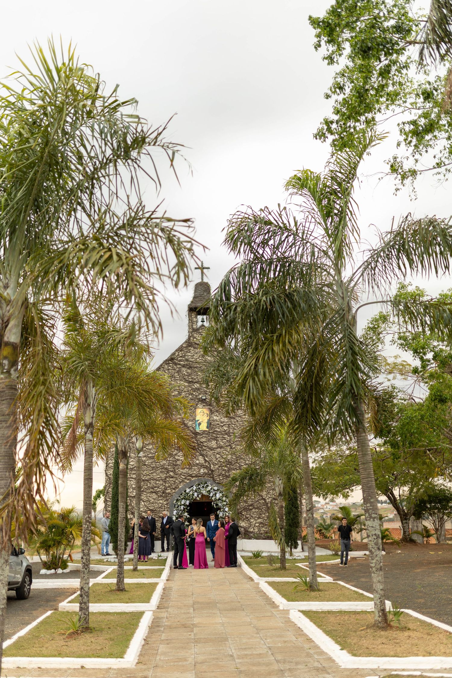 Stone church with bell tower surrounded by palm trees, with wedding guests gathered at the entrance along a paved pathway.