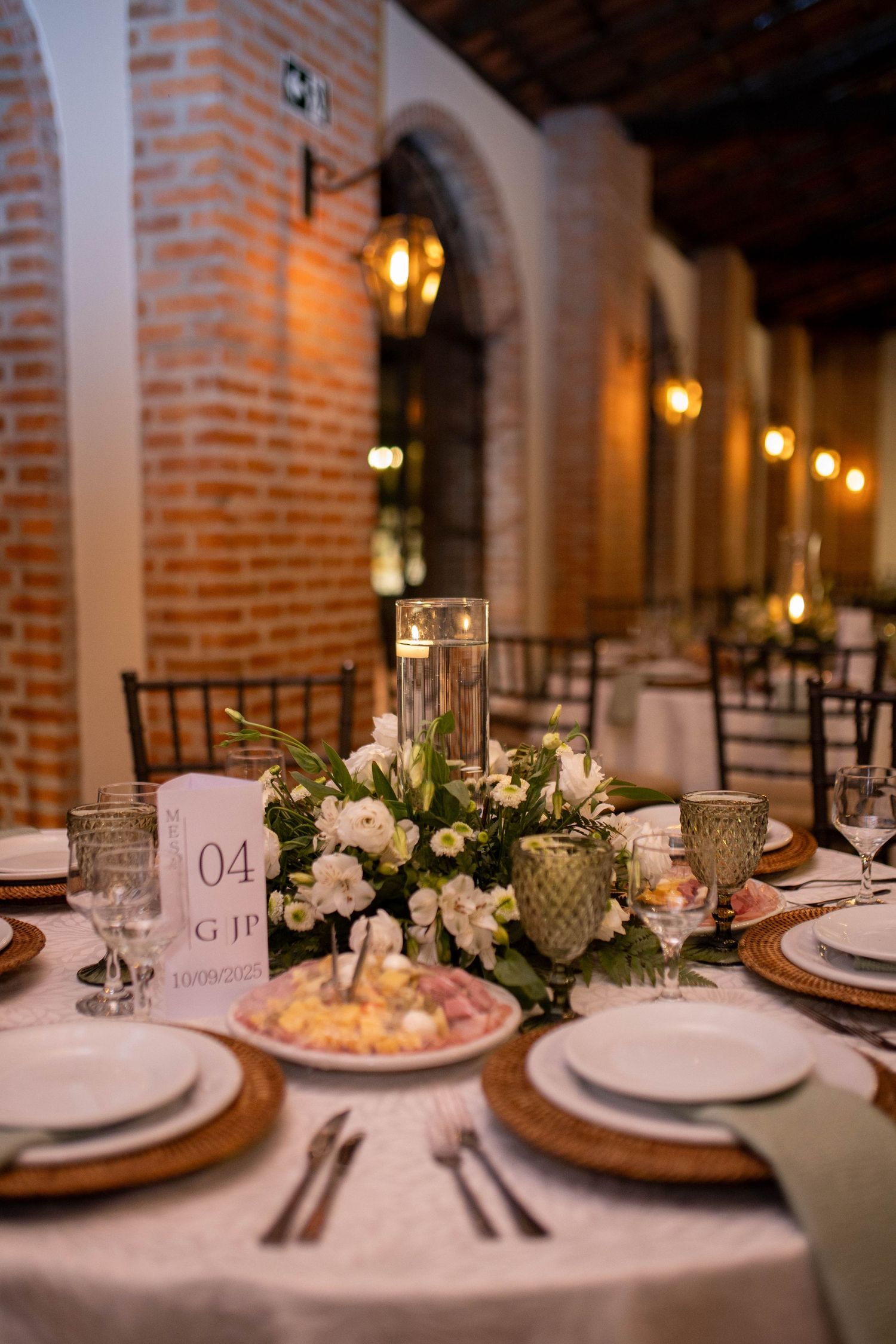 Elegant wedding reception table with white floral centerpiece, candles, gold chargers, and glassware in a brick venue.