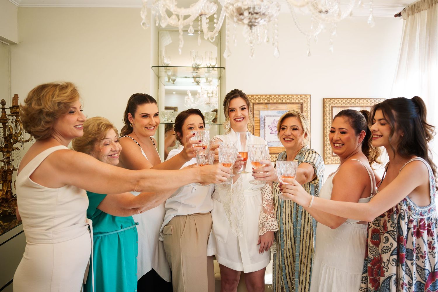 A bride in white robe toasts champagne with bridesmaids and family in an elegant room with chandelier.
