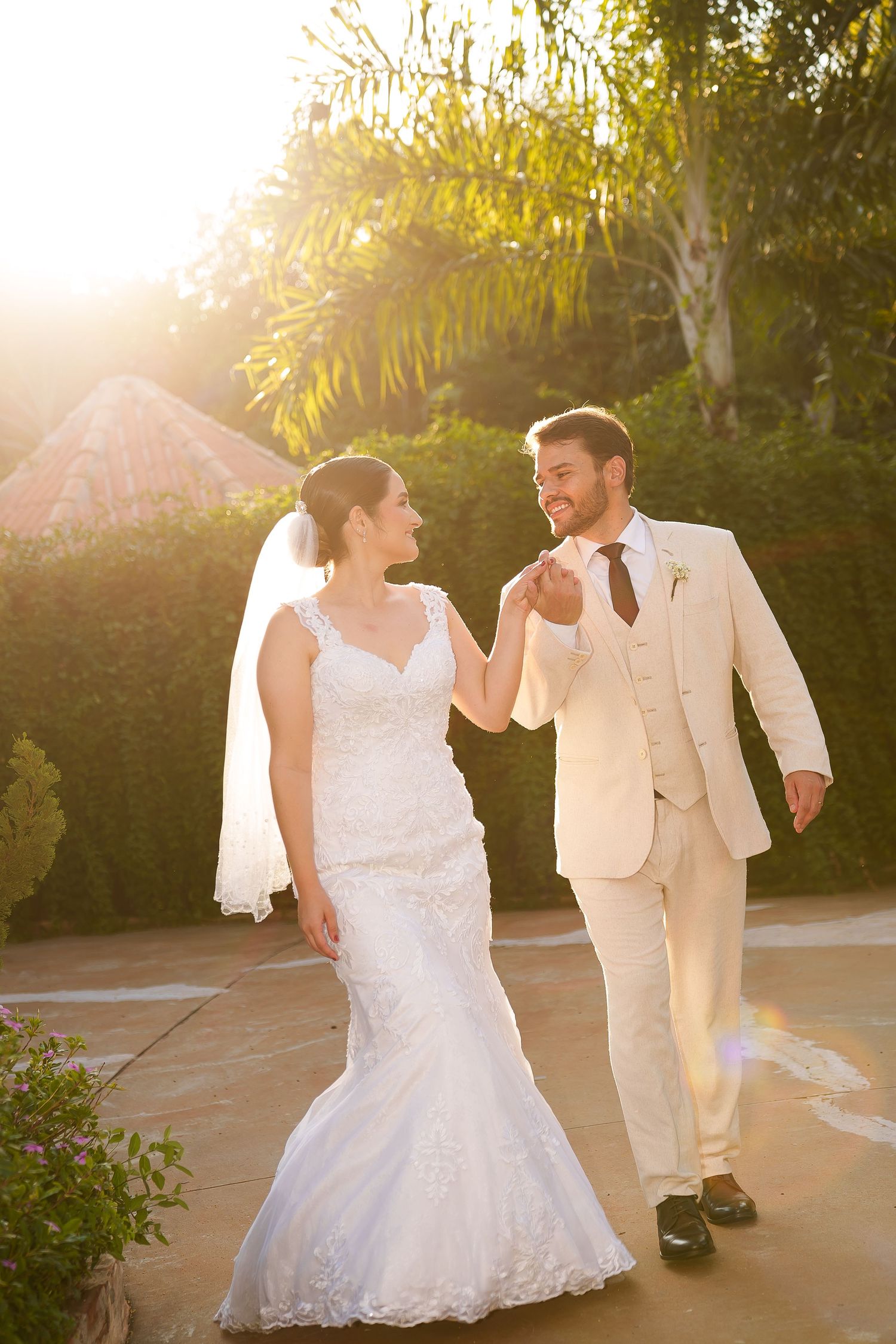 Bride and groom walking together outdoors in golden sunlight, she in lace gown with veil, he in cream suit.