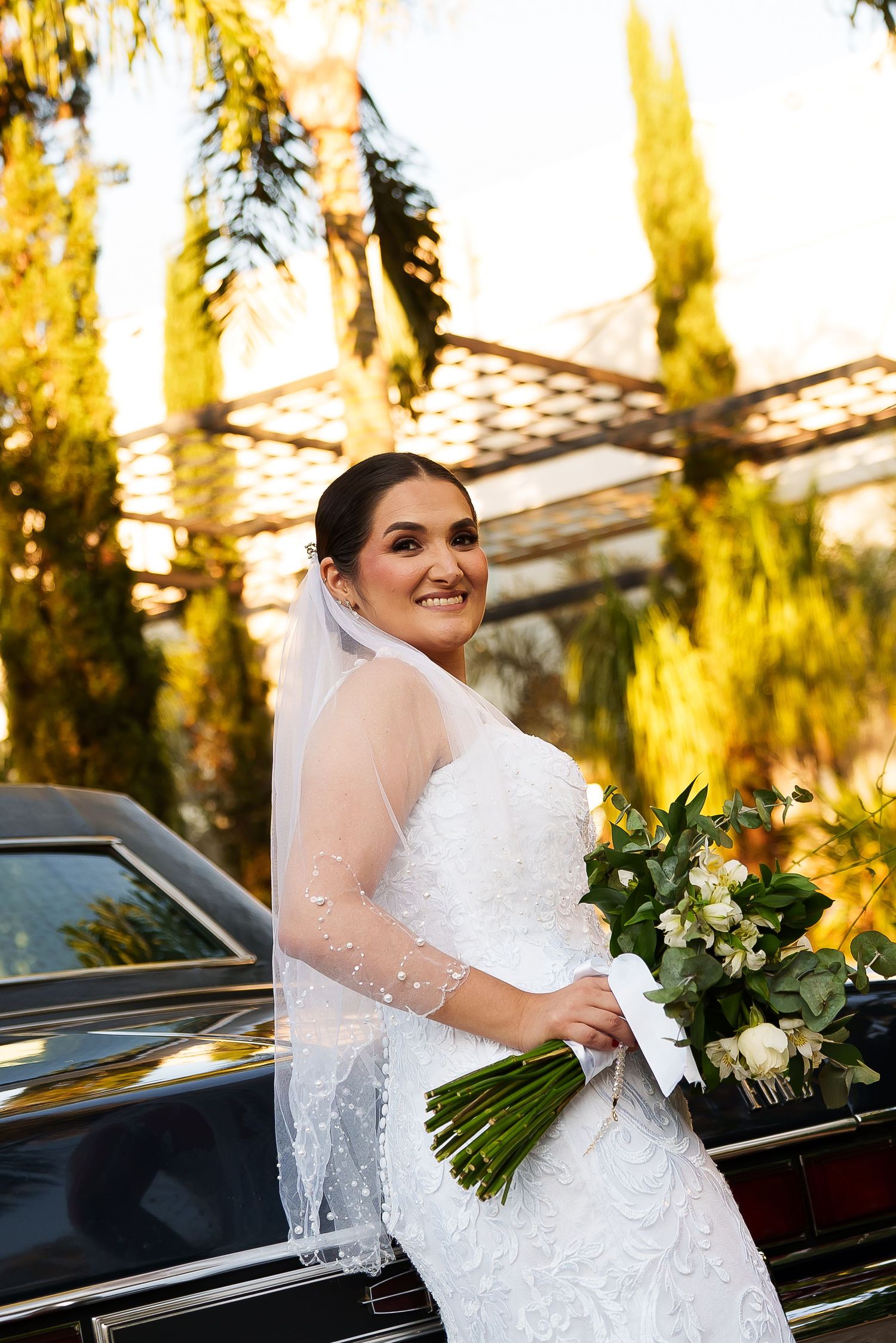 Smiling bride in lace gown and pearl veil holds white bouquet beside classic black car surrounded by golden palm trees.