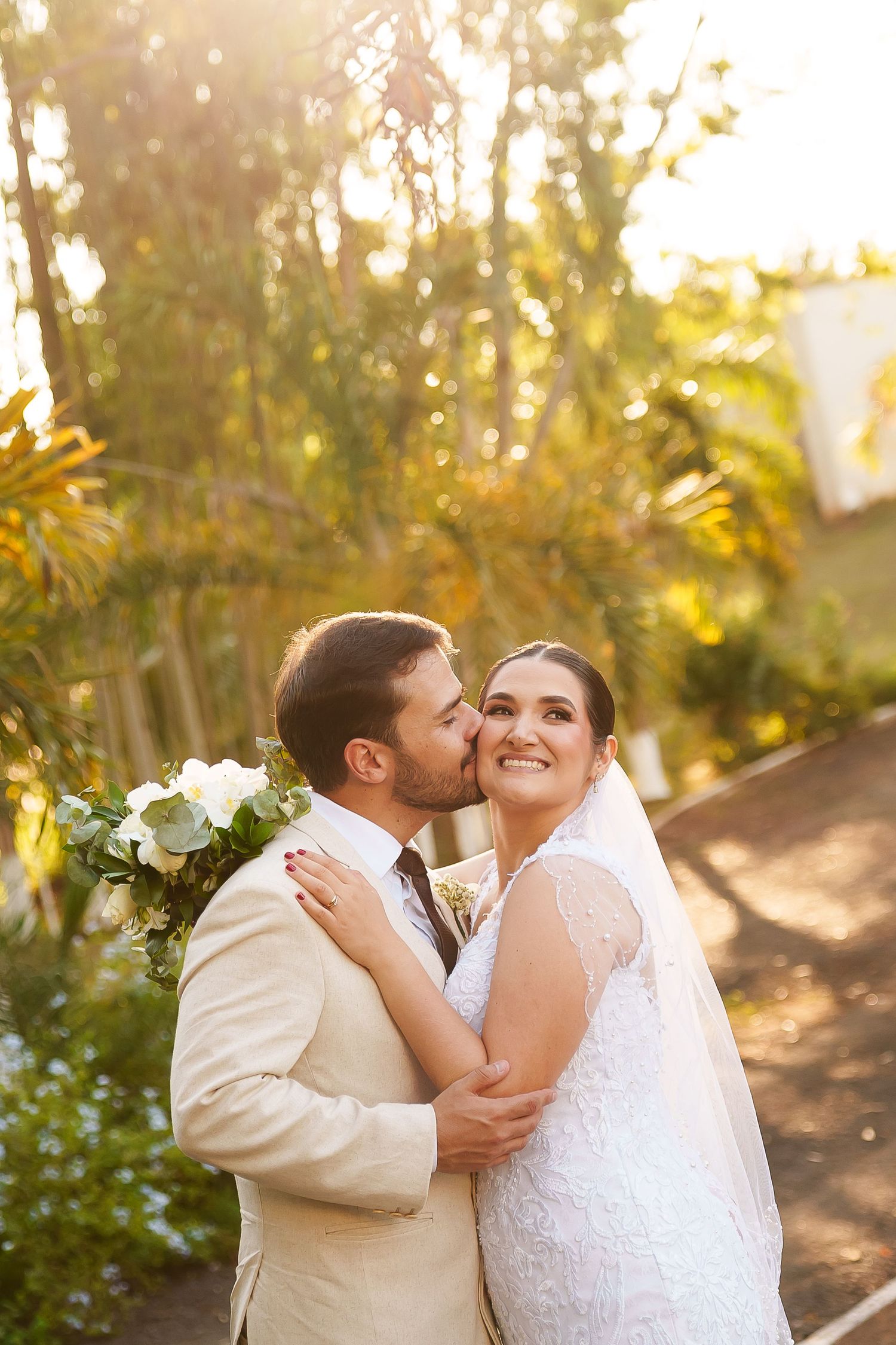 Groom in beige suit kisses bride in white lace gown and veil, holding white flowers, in a sunlit garden setting.
