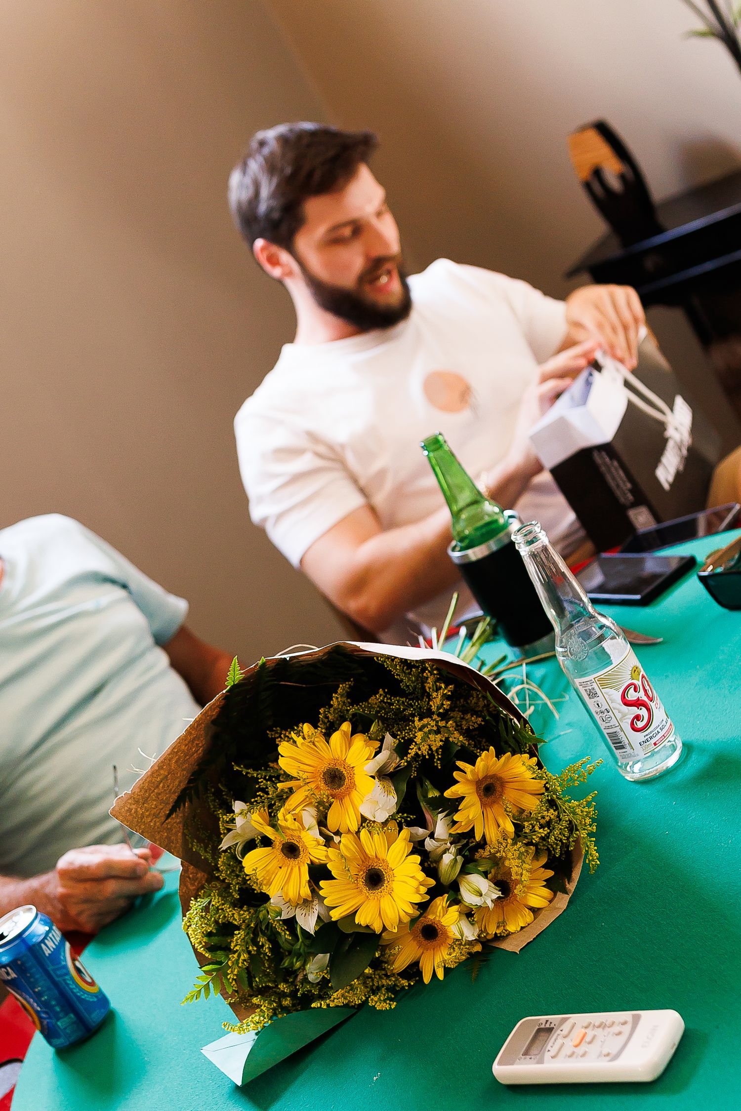 A bouquet of yellow gerbera daisies on a green table with bottles and a bearded man opening a gift in the background.