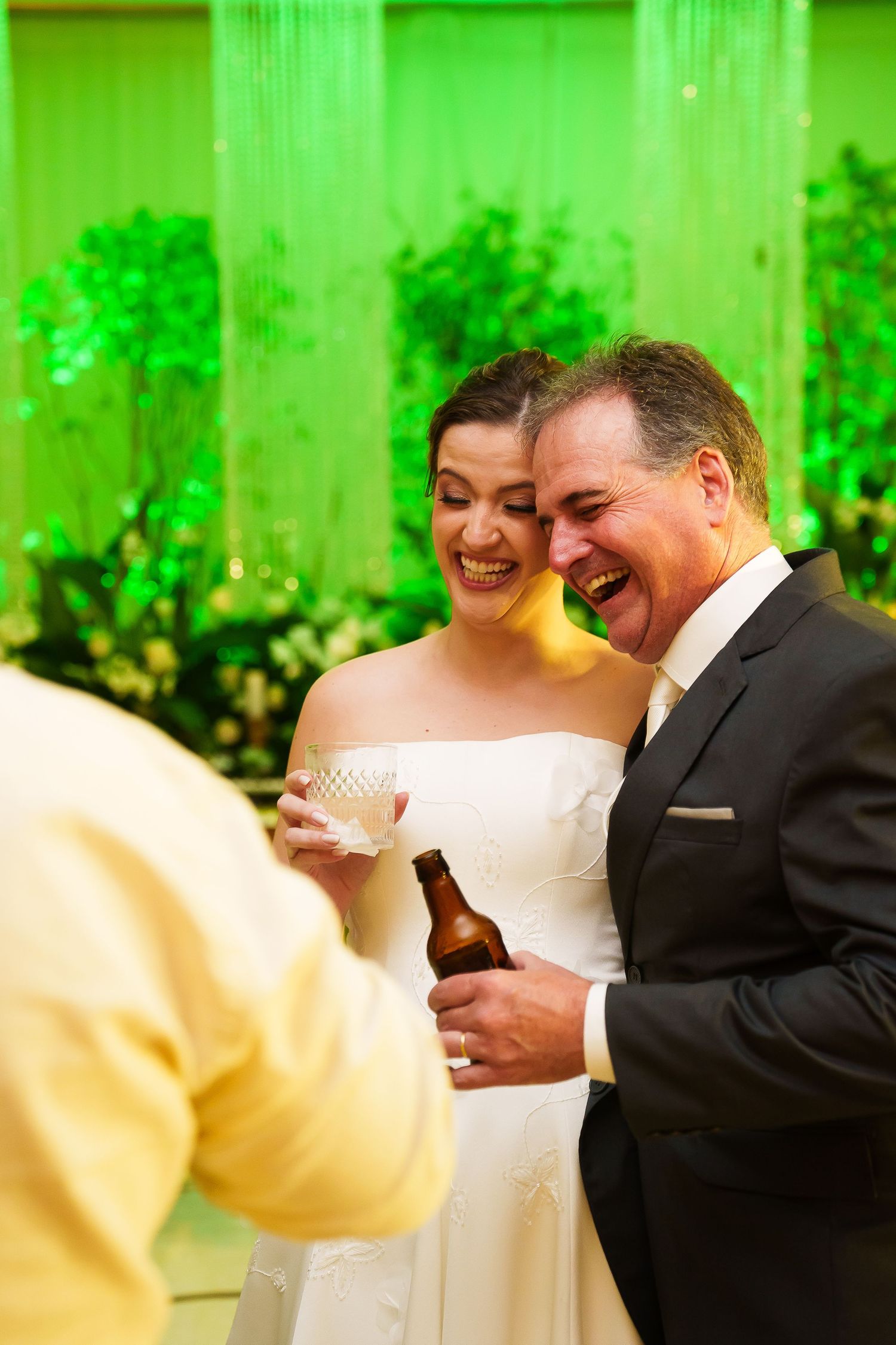 Bride and groom laughing together at wedding reception, holding drinks with green lit backdrop.