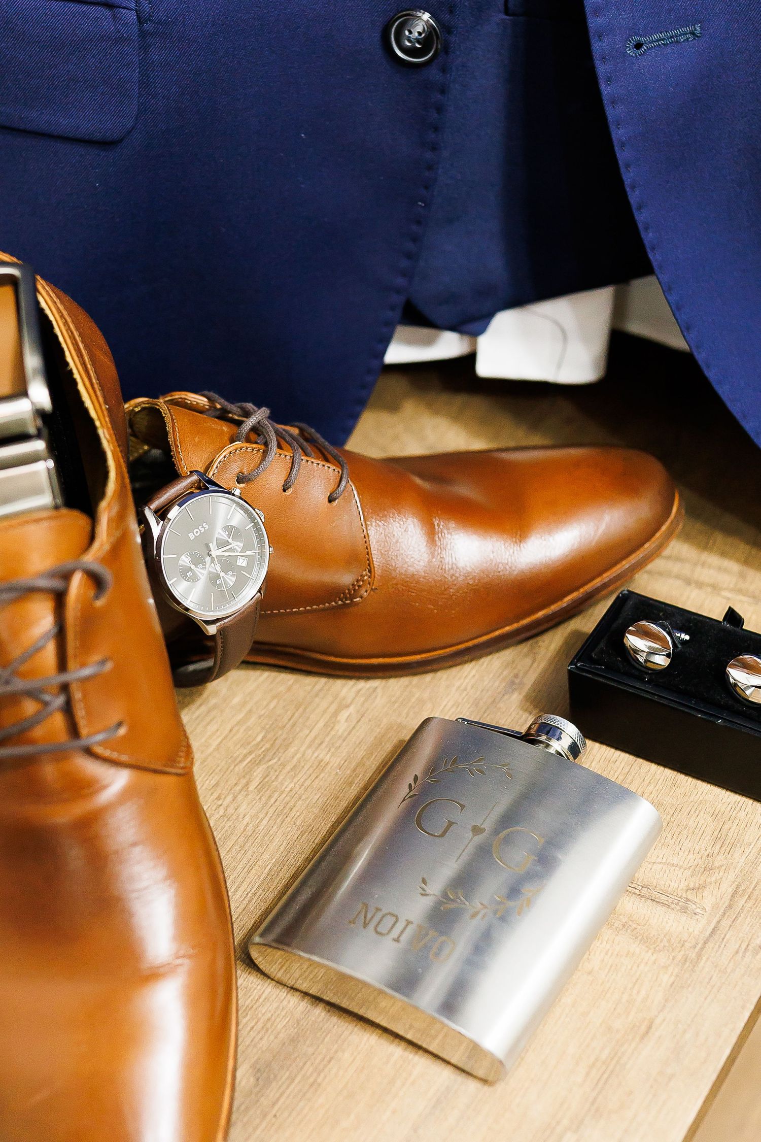 Brown leather dress shoes, silver flask, watch, and cufflinks arranged on wooden floor beside a blue suit for a wedding.