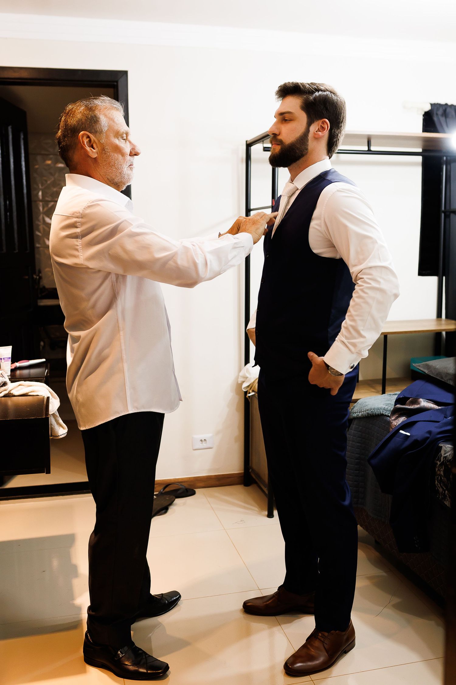 Older man helping young groom adjust his tie while getting dressed in a hotel room before wedding.