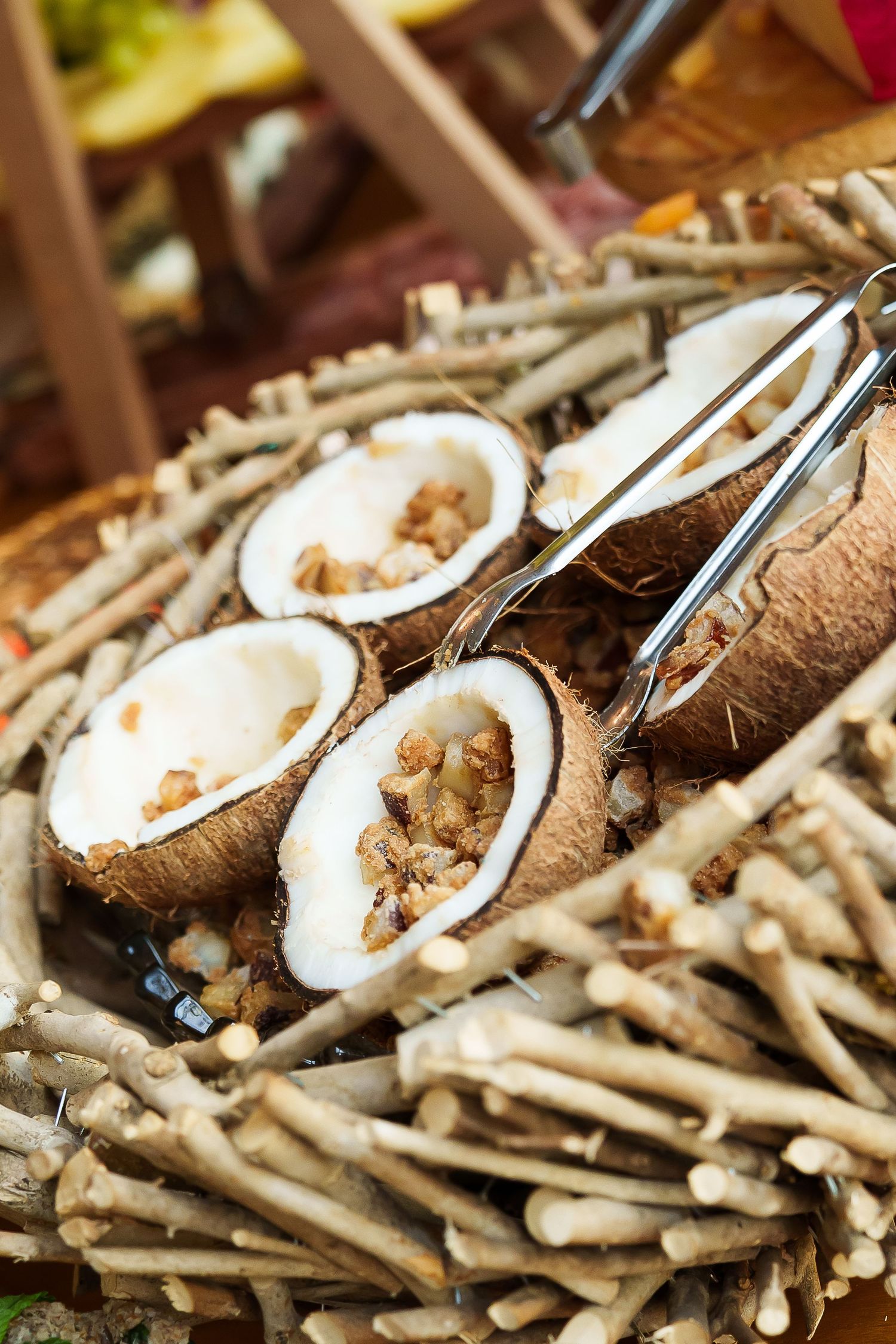 Halved coconuts filled with nuts displayed on a rustic twig basket with serving tongs at an outdoor food market.