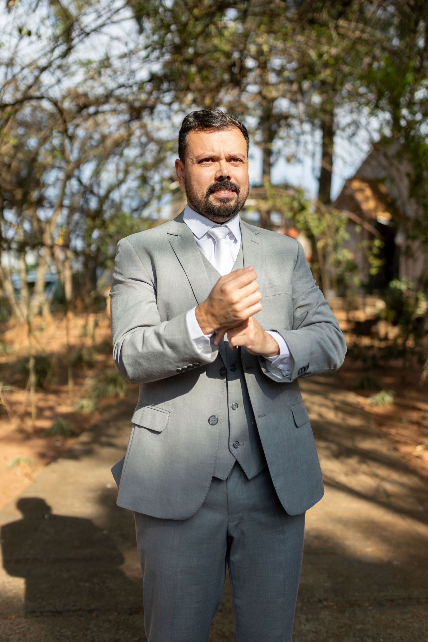 Man in gray three-piece suit adjusting cufflinks outdoors among trees on a sunny day.