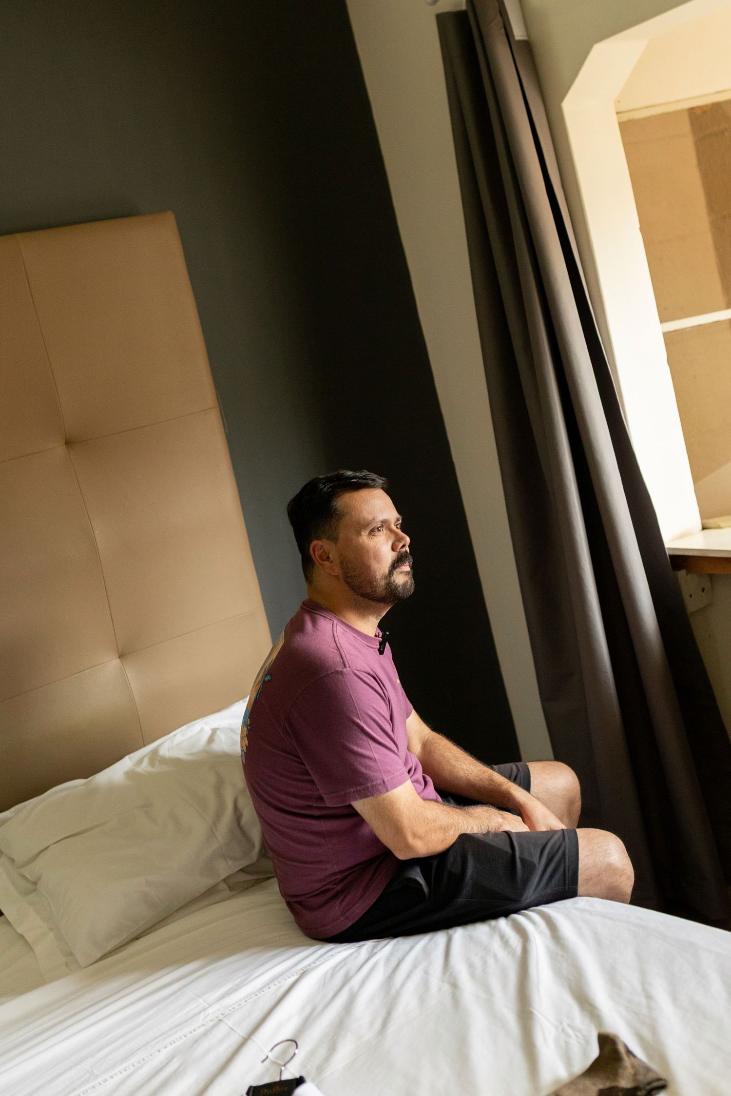 Man in purple shirt sitting cross-legged on hotel bed, gazing thoughtfully toward window with curtains.