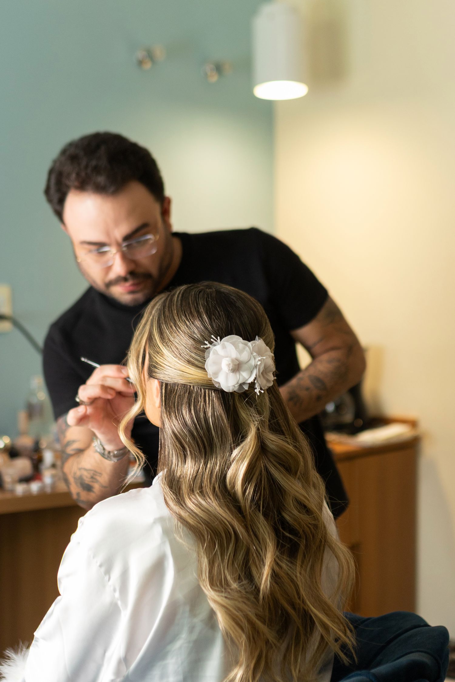 Makeup artist applying makeup to a bride with wavy hair adorned with a white flower clip in a salon.