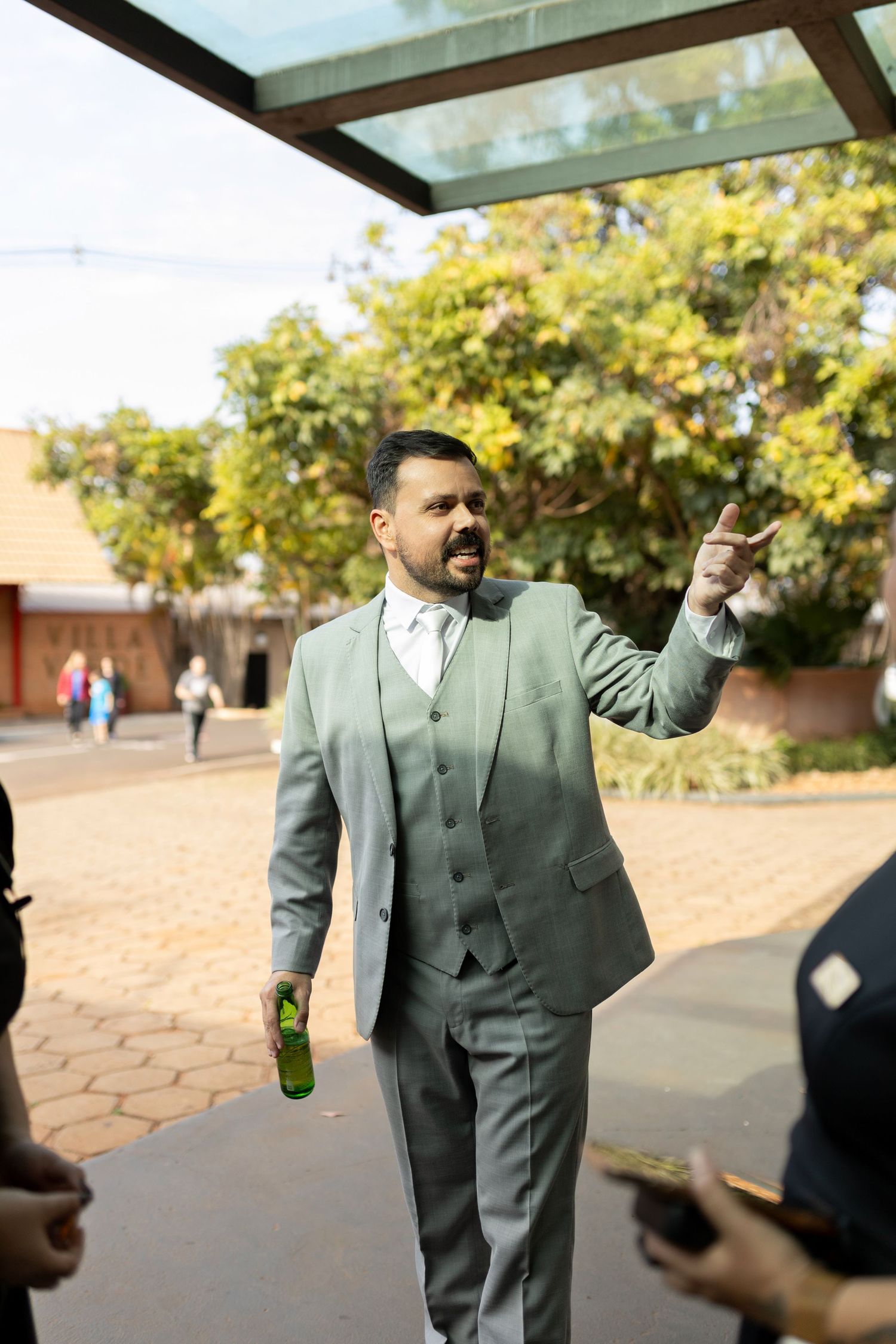 Man in gray suit holding green bottle and pointing outdoors near glass canopy with trees in background.