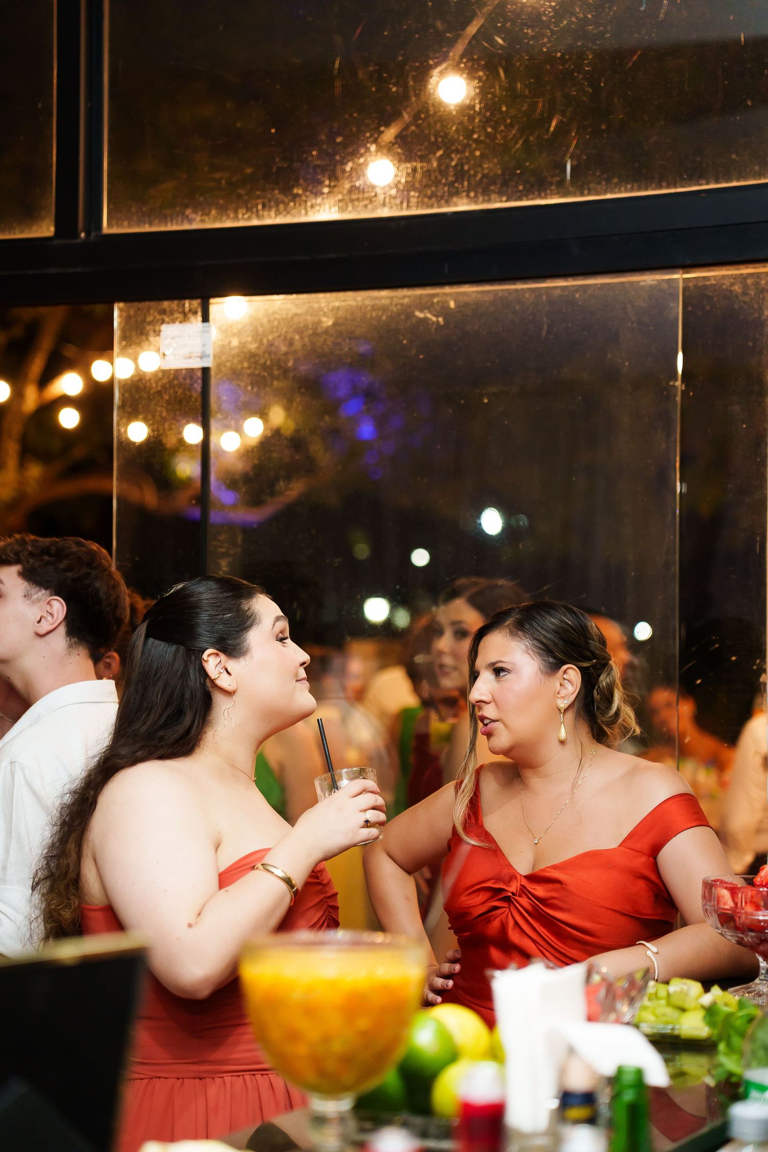 Two women in red dresses chat at an elegant evening event, with string lights and a fruit-laden table in the foreground.