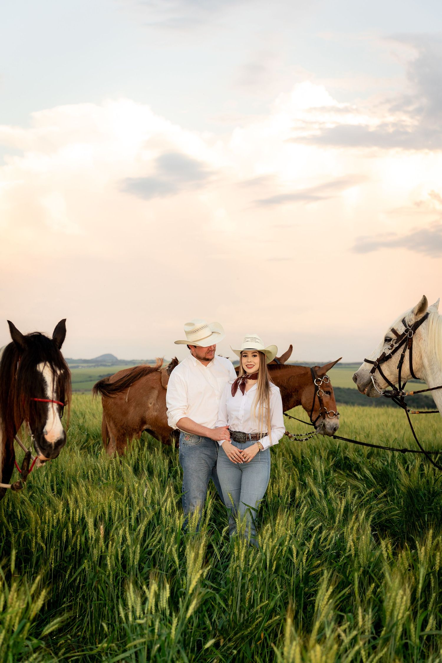 Couple in cowboy hats standing together in a green field at sunset, surrounded by brown and white horses.