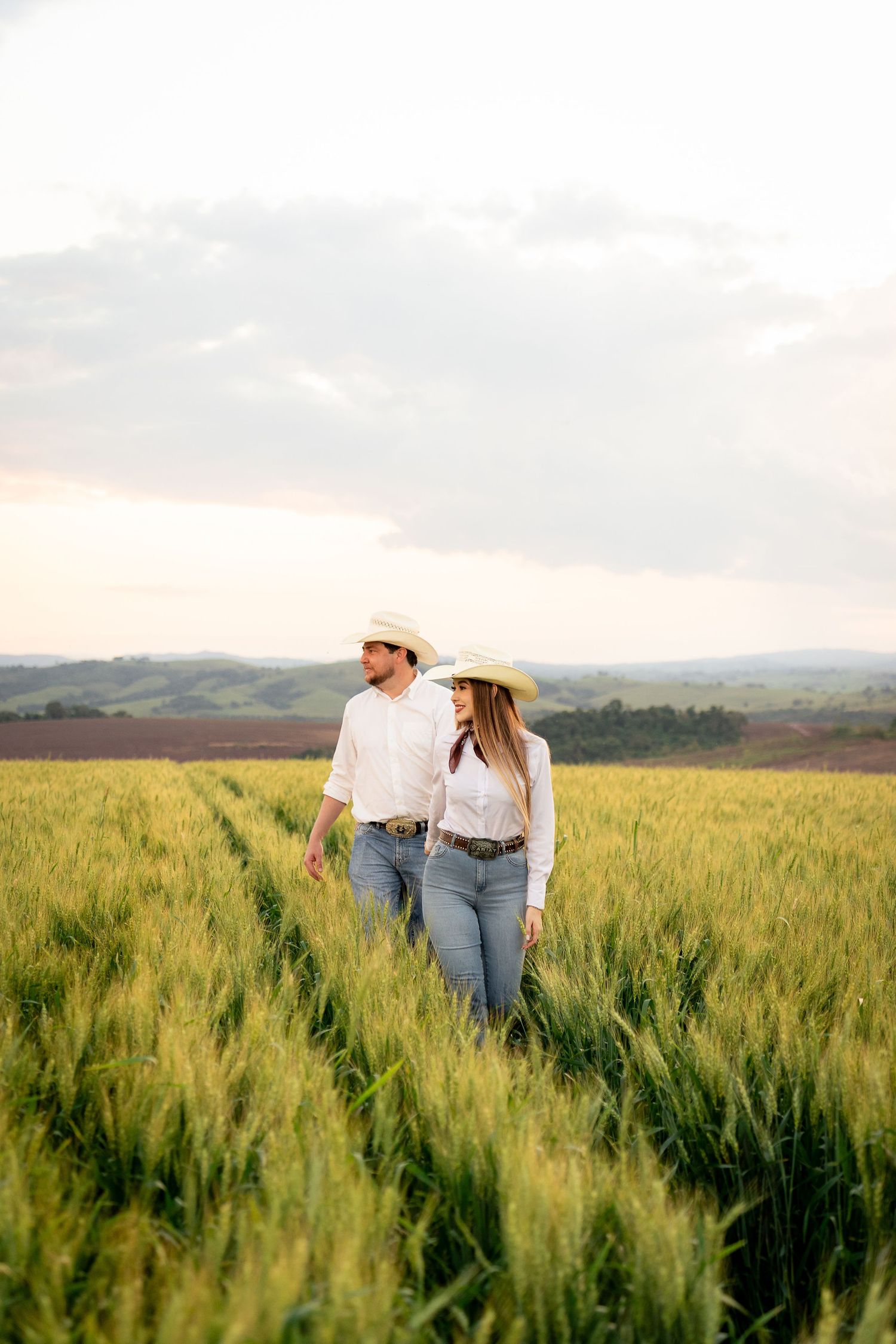 Couple in cowboy hats and jeans walking through a lush green wheat field at sunset with rolling hills behind.