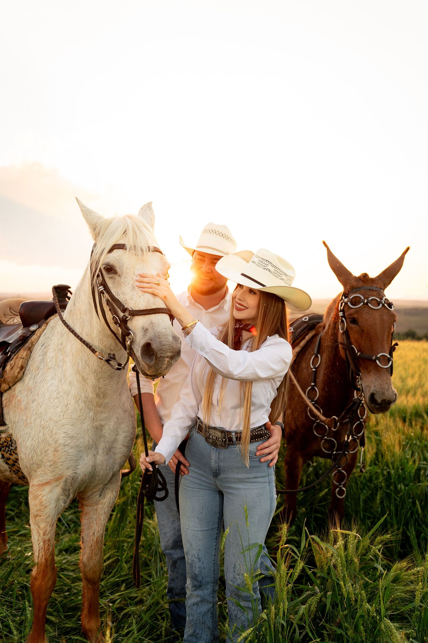 Two cowboys in white hats stand with a white and brown horse in a sunlit green field at golden hour.