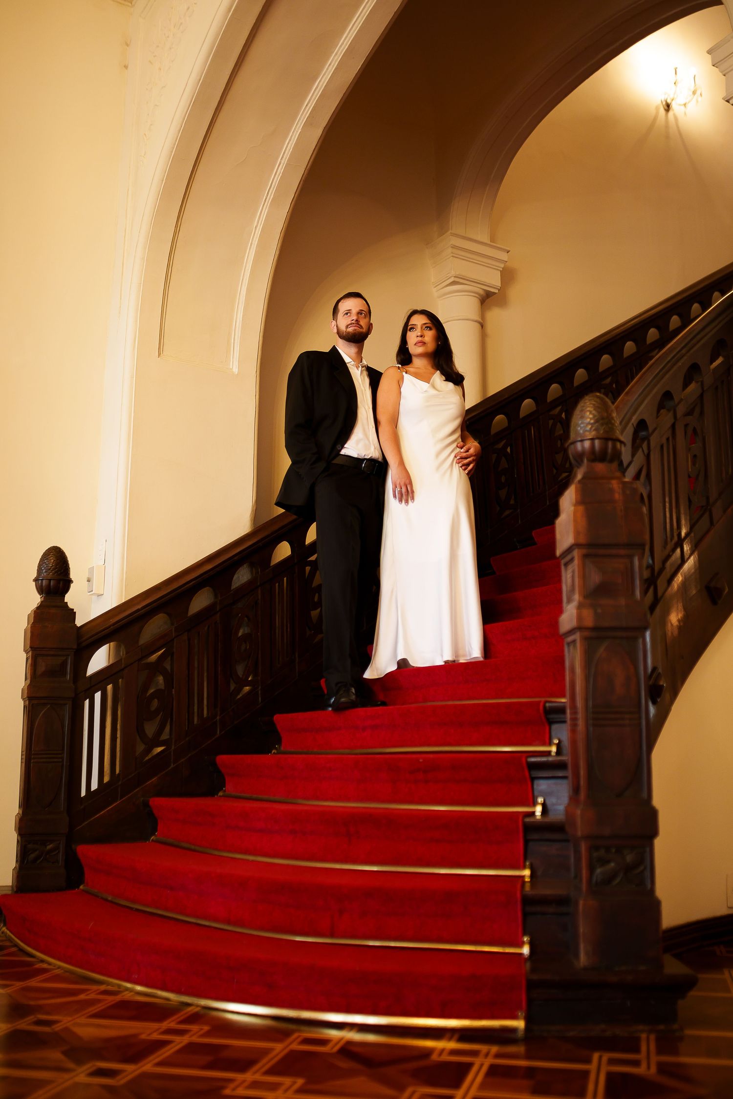 Elegant couple in formal attire posing on grand staircase with red carpet and dark wood railings.