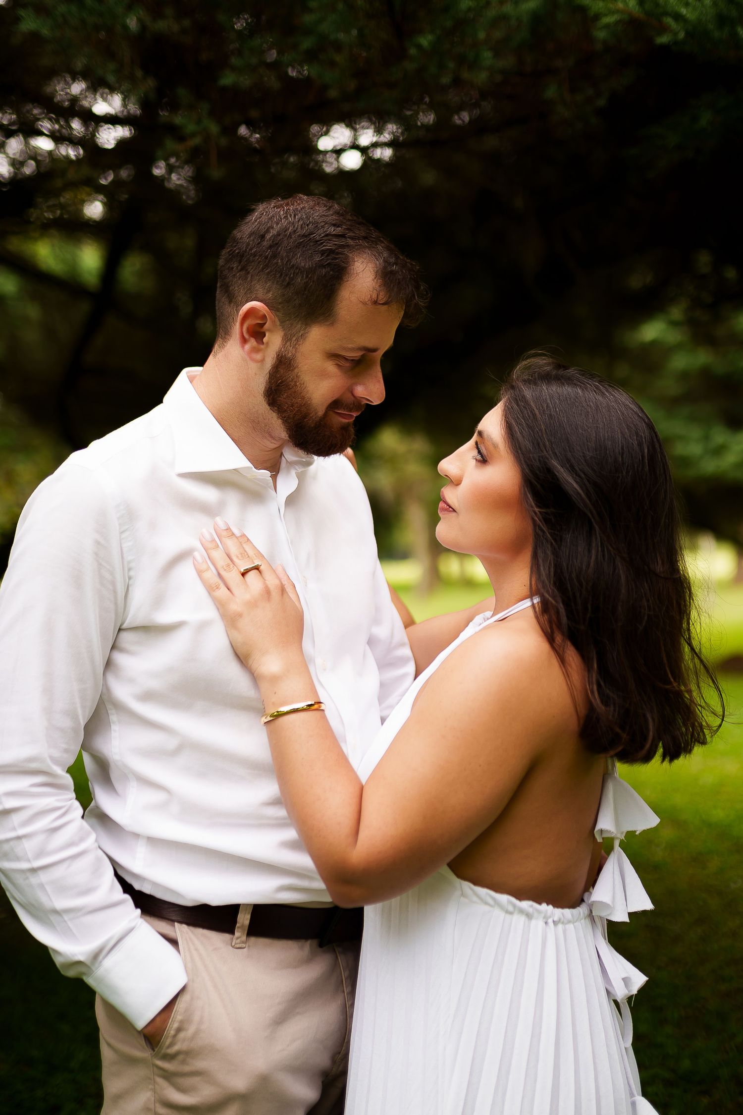 Couple in white outfits sharing a romantic moment outdoors, woman wearing engagement ring, green trees in background.