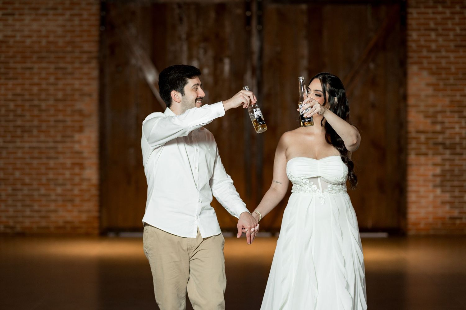 Bride and groom toasting with drinks in a rustic barn venue, holding hands in wedding attire.