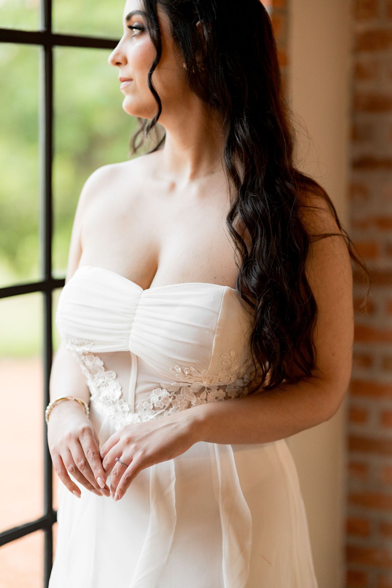 Bride in strapless white wedding gown with floral lace detail stands by window with long dark wavy hair.